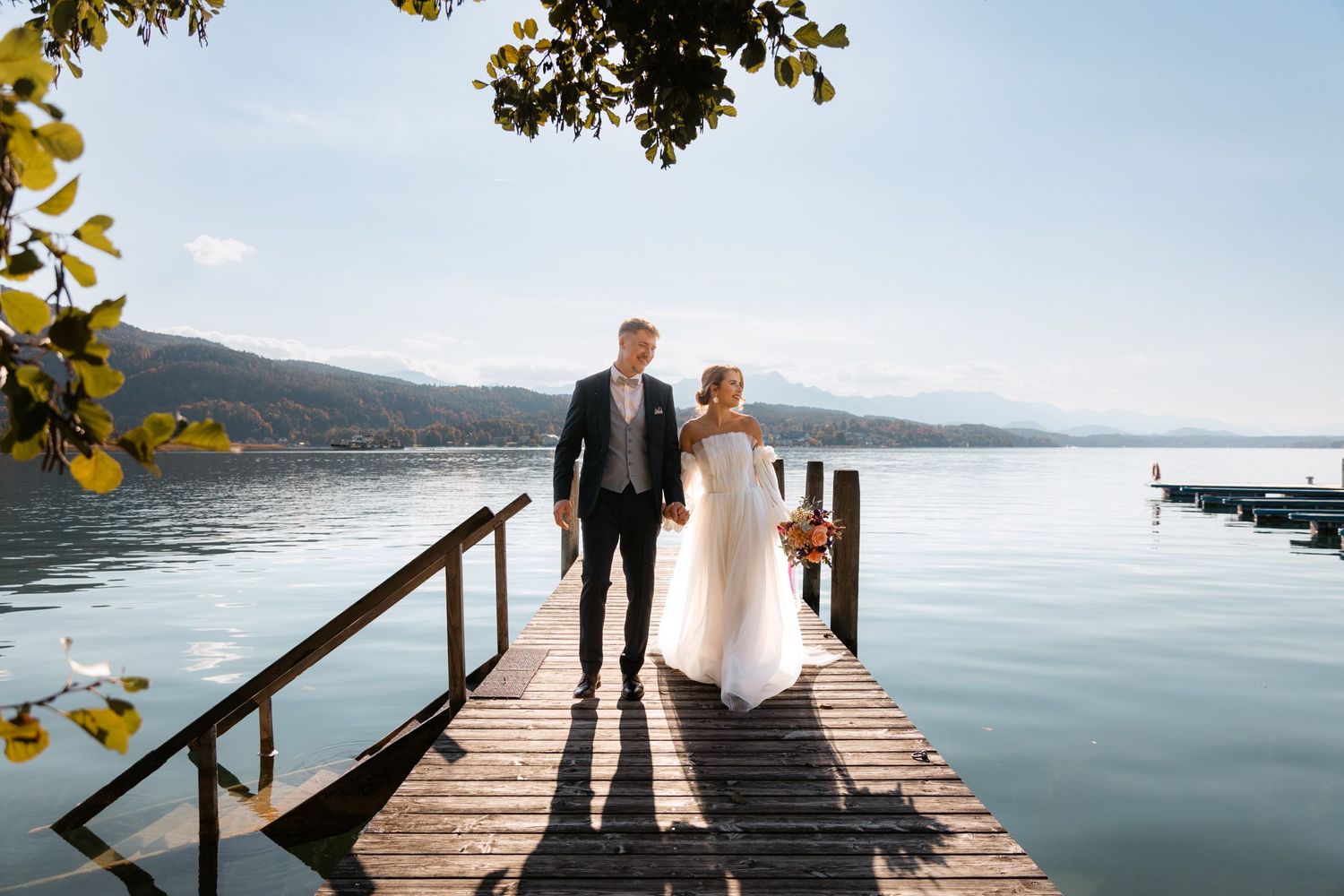 Bride and groom walking hand in hand on a wooden dock overlooking a serene mountain lake.