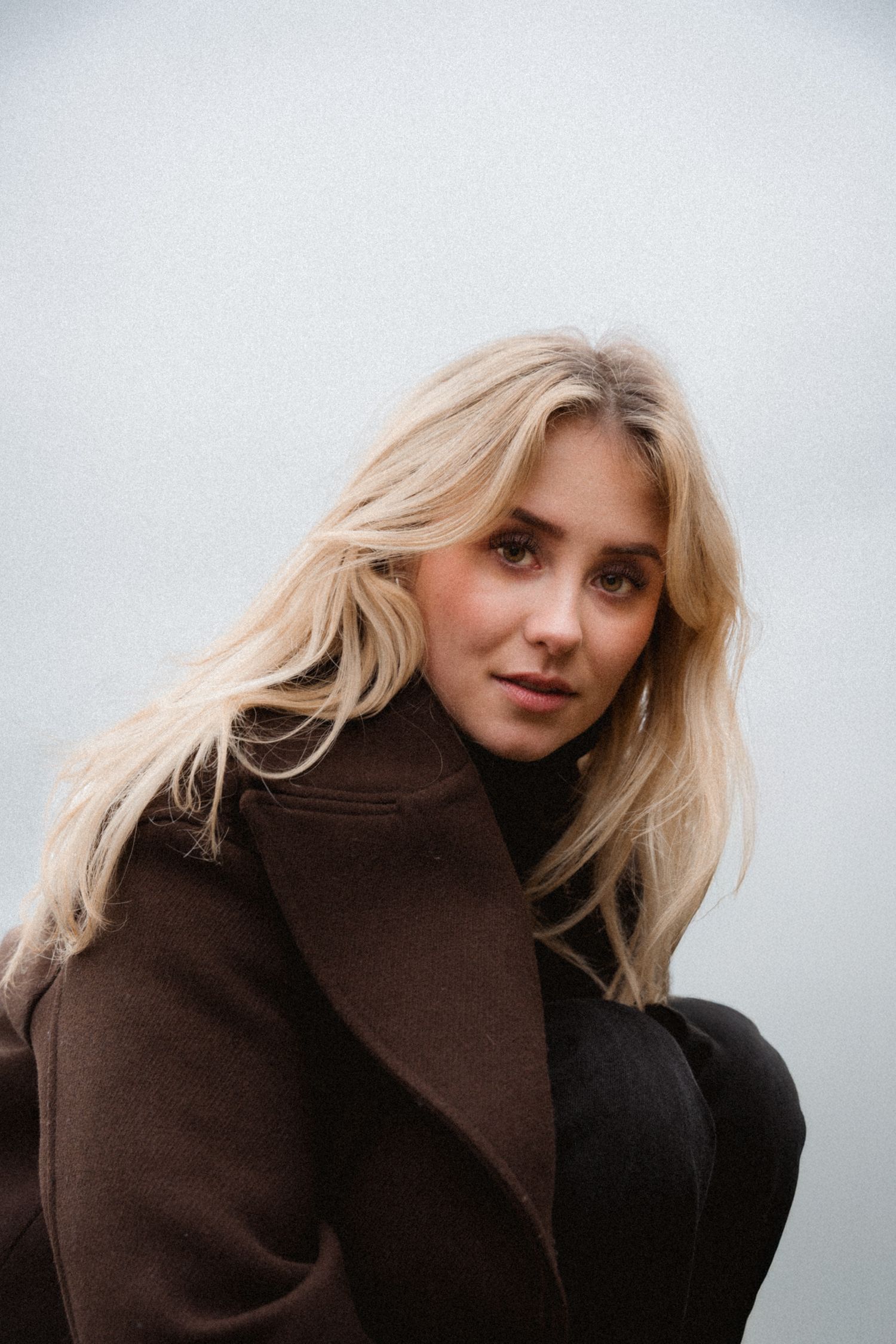 Blonde woman in brown coat posing outdoors against grey background.