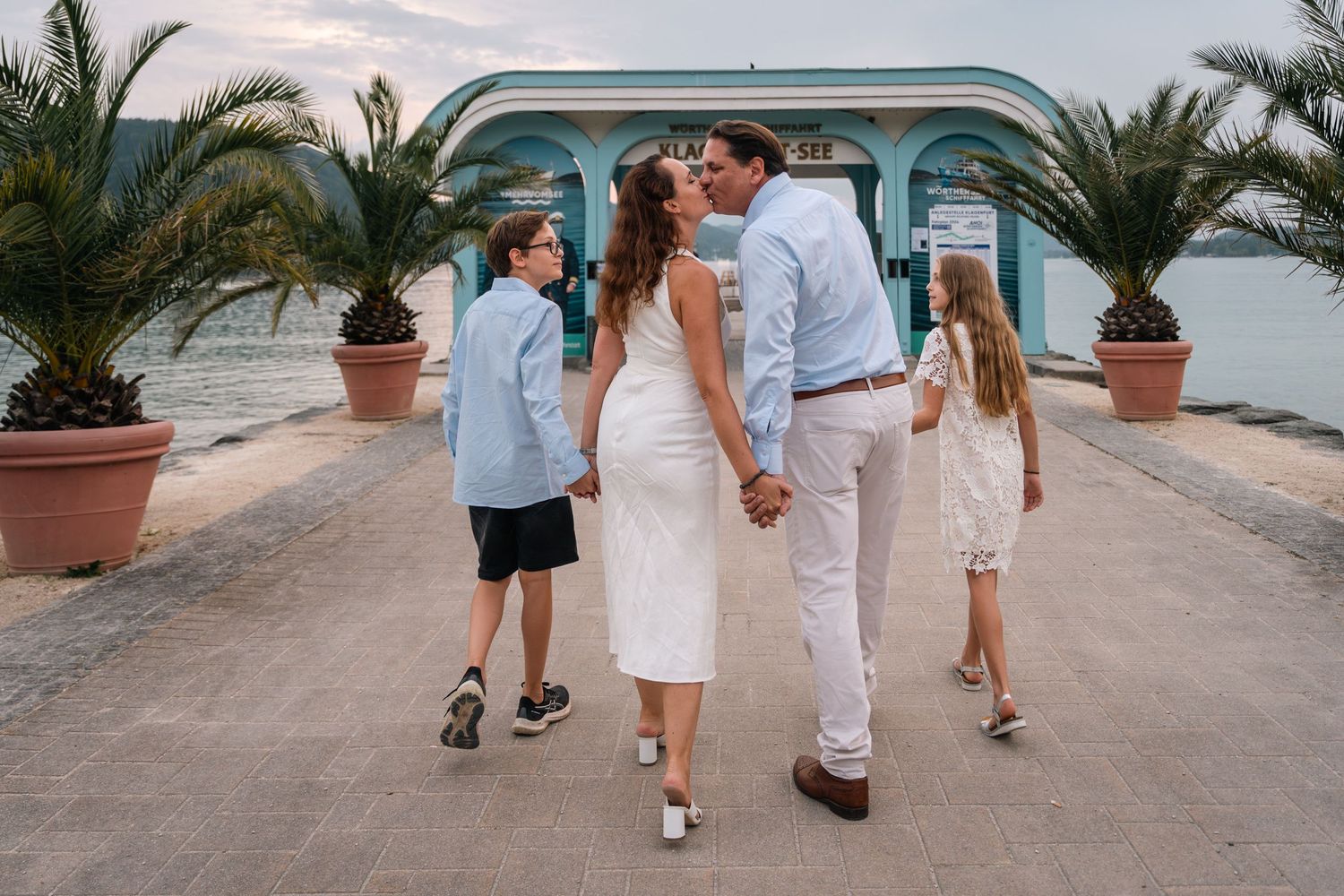 Family of four walking hand-in-hand toward a waterfront pavilion, couple kissing while children walk beside them.