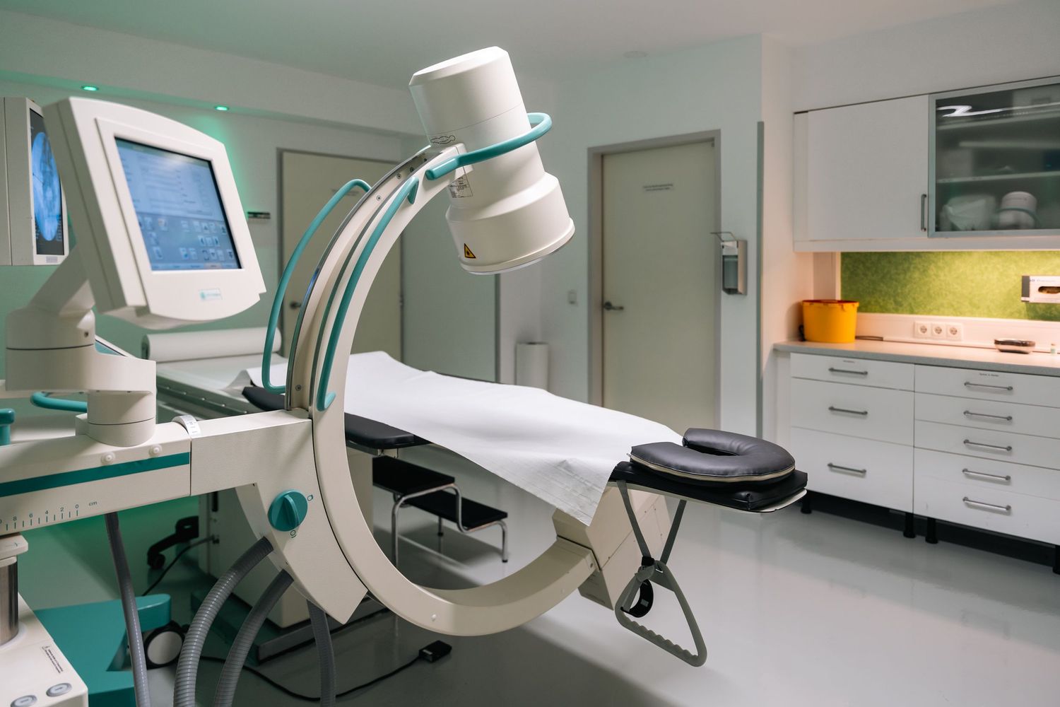 Medical imaging room with C-arm fluoroscopy machine and examination table in clinical setting.