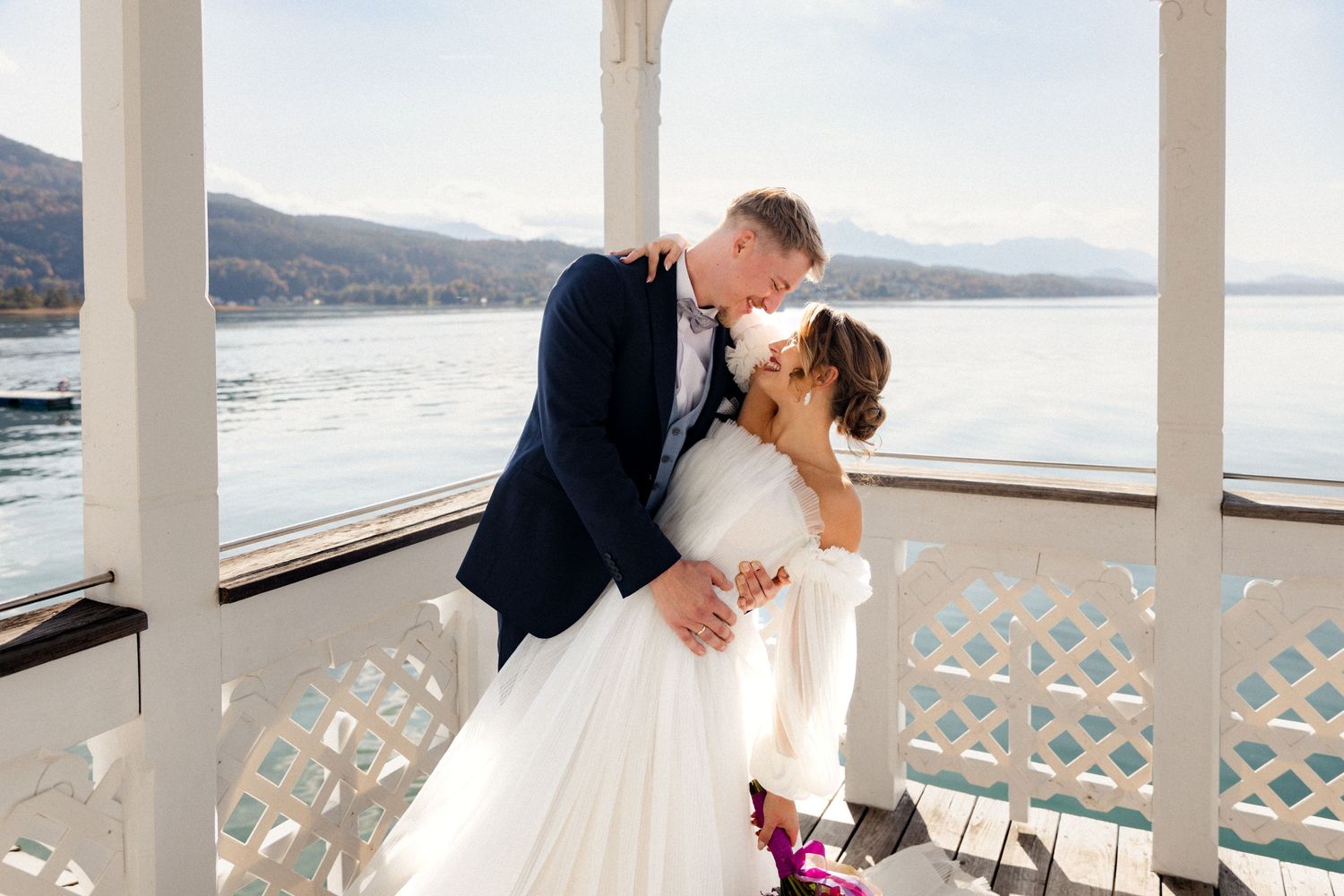 Bride and groom sharing a romantic kiss on a lakeside pavilion with mountain views.