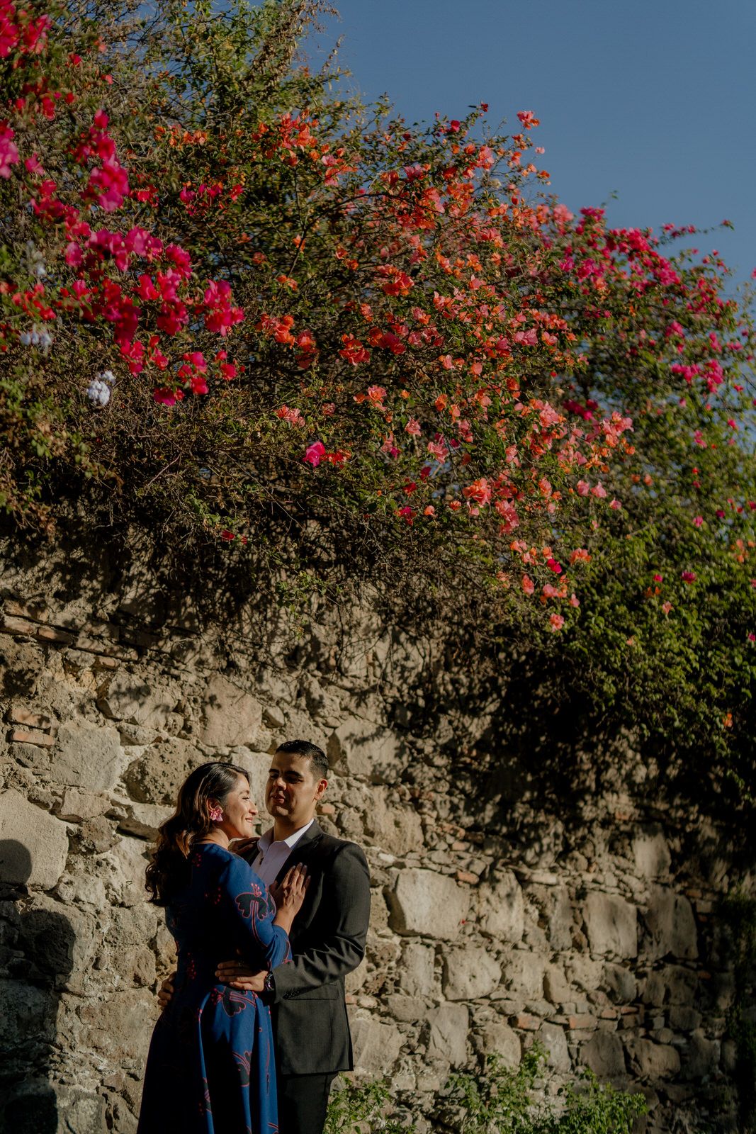 A tender embrace beneath flowering bougainvillea and a rustic stone wall.