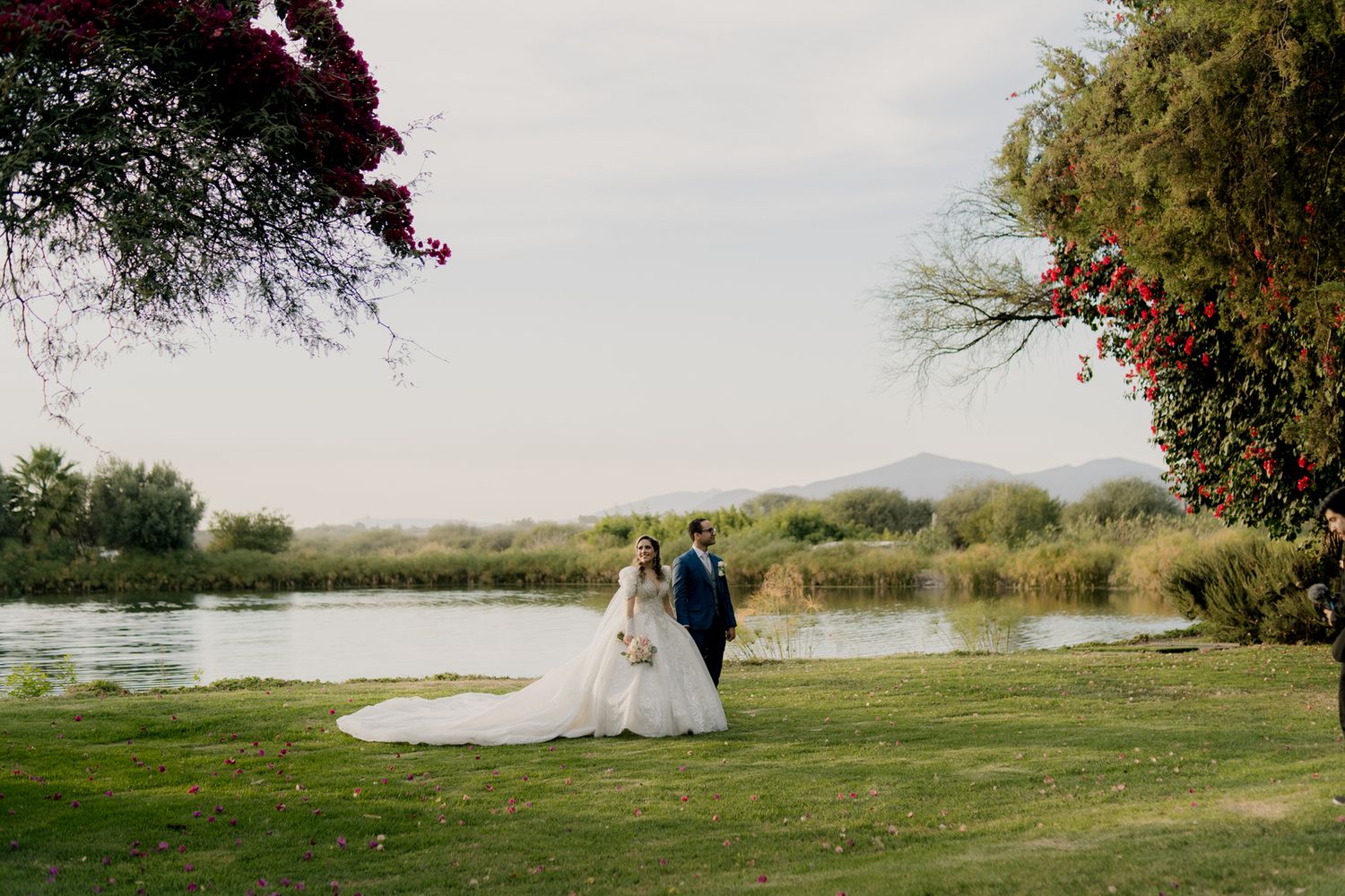 A couple stands by a serene lake with mountains and flowering trees in the background.