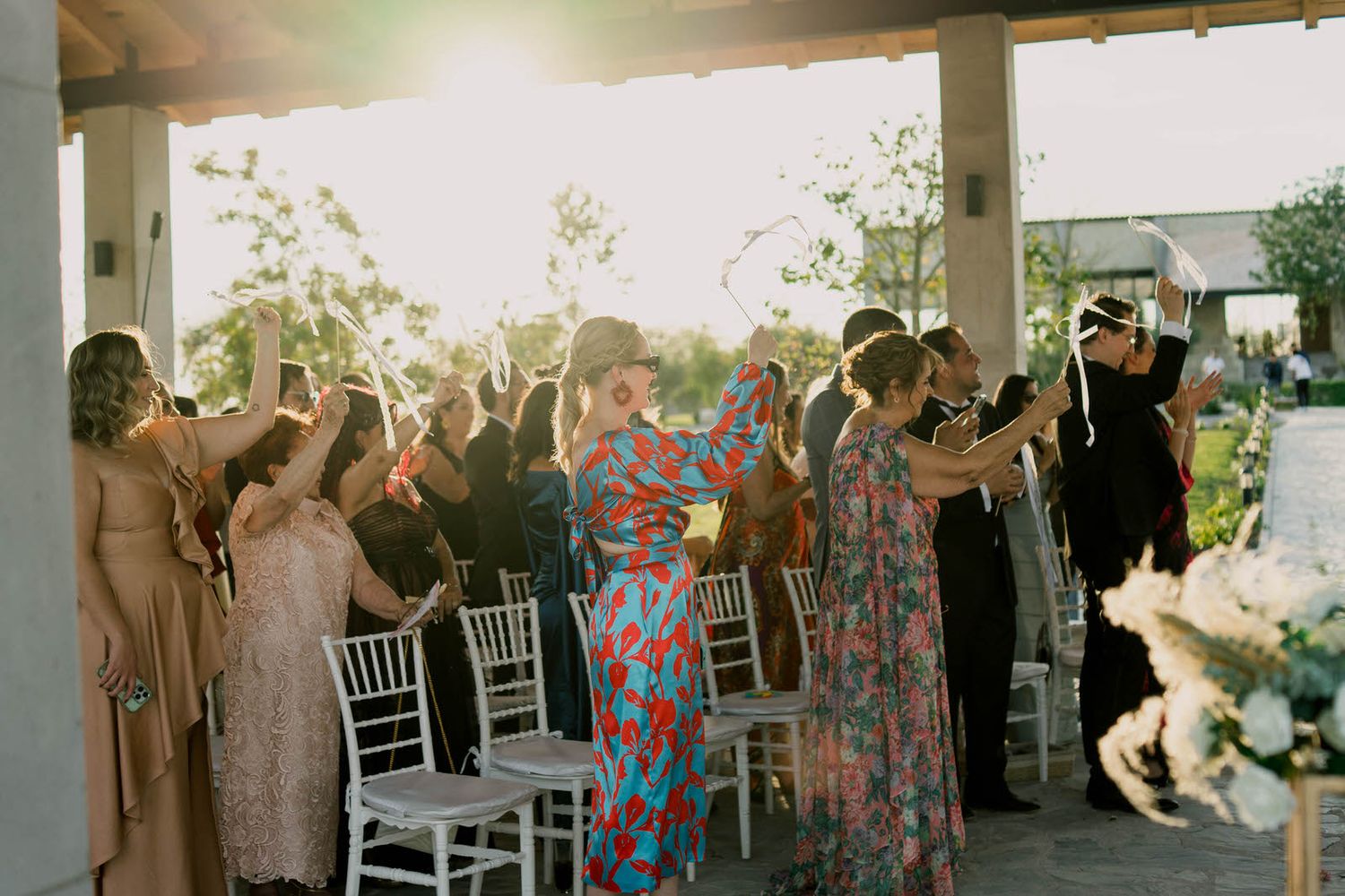 Wedding guests in colorful dresses celebrate with raised arms under an outdoor pavilion.