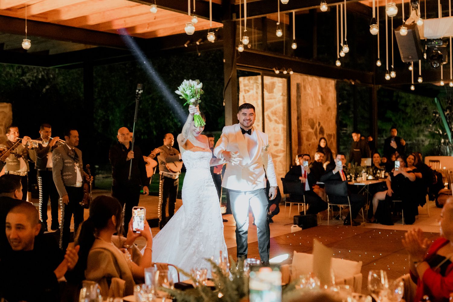 Warm-lit reception venue with hanging lights and dancing guests in the background.