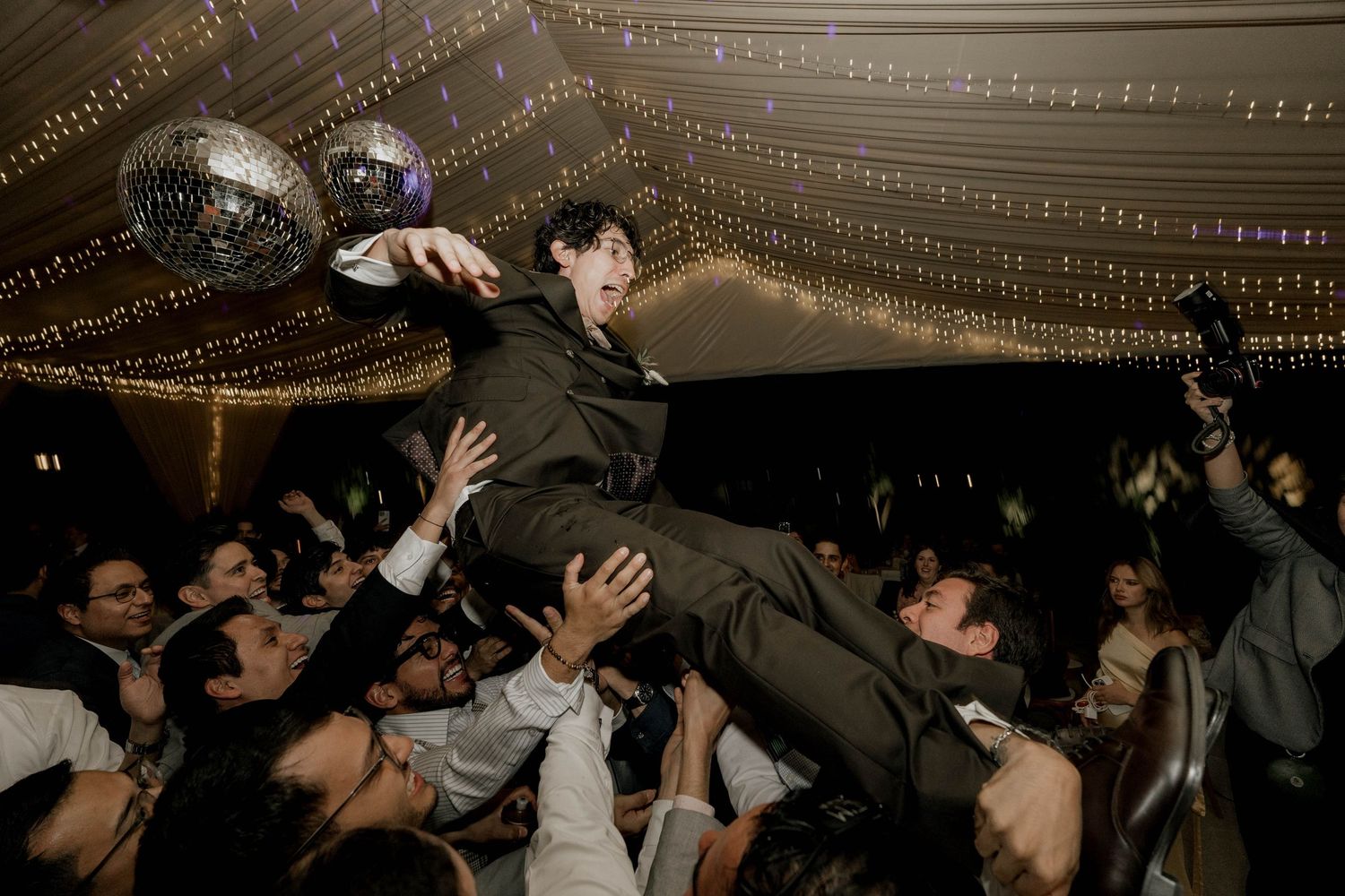 Wedding guests dancing in a dimly lit reception tent with string lights overhead.