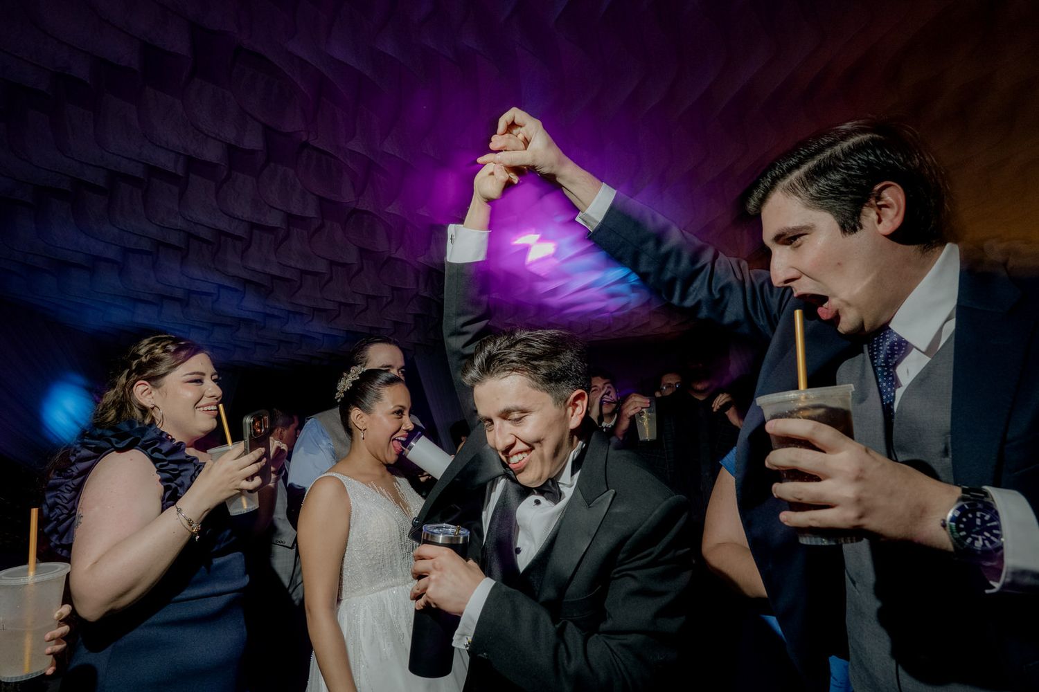Bride and groom celebrate with guests during lively wedding reception party under purple lights.