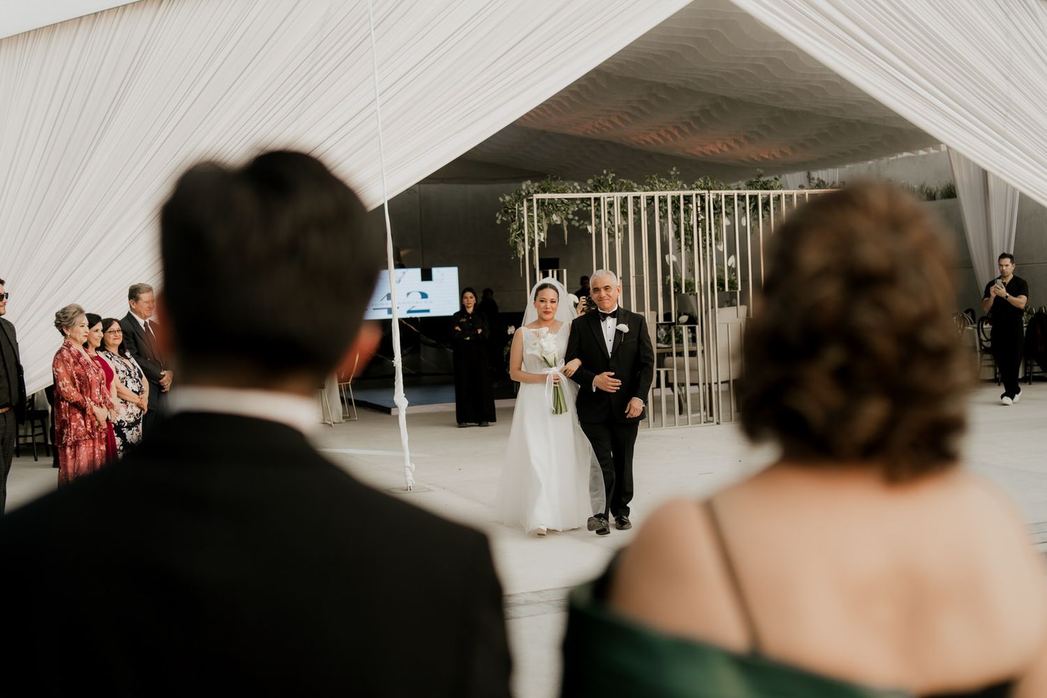Bride and escort walk toward groom at outdoor wedding ceremony under white tent with seated guests.