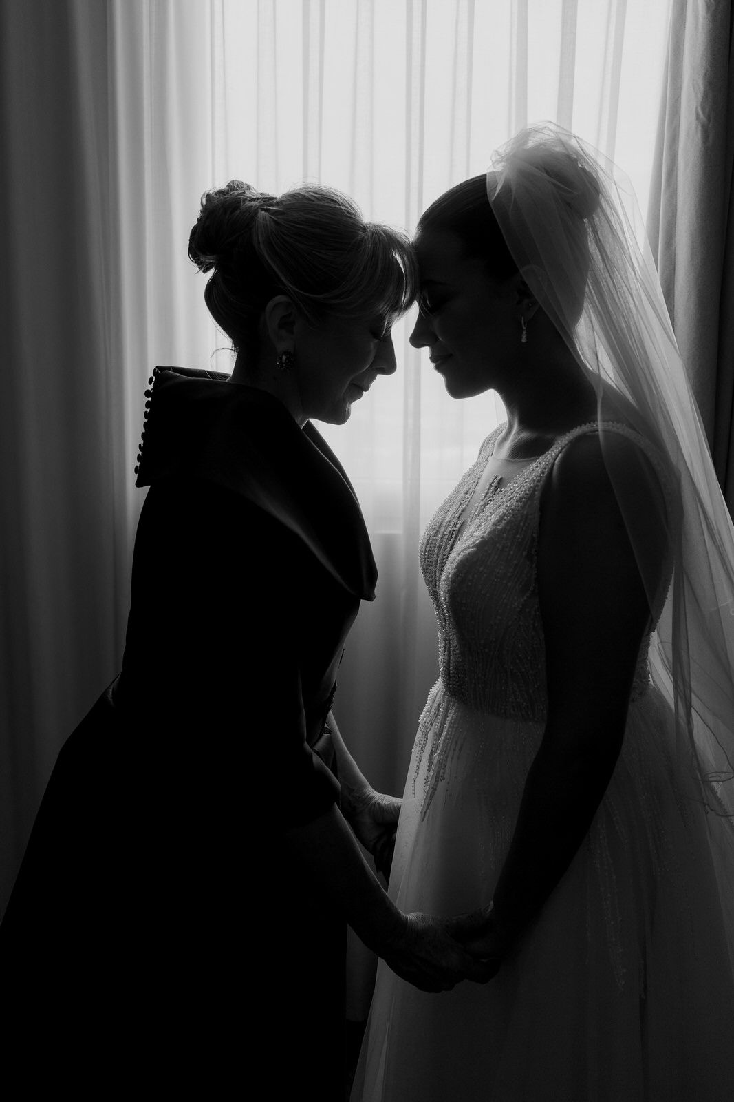 Black and white silhouette of bride in veil and woman holding hands by window.
