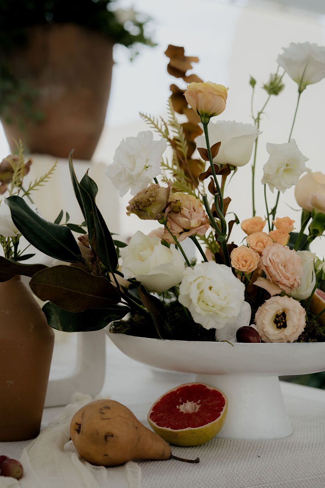 Elegant floral centerpiece with white and peach roses beside a halved grapefruit and pear on a table.