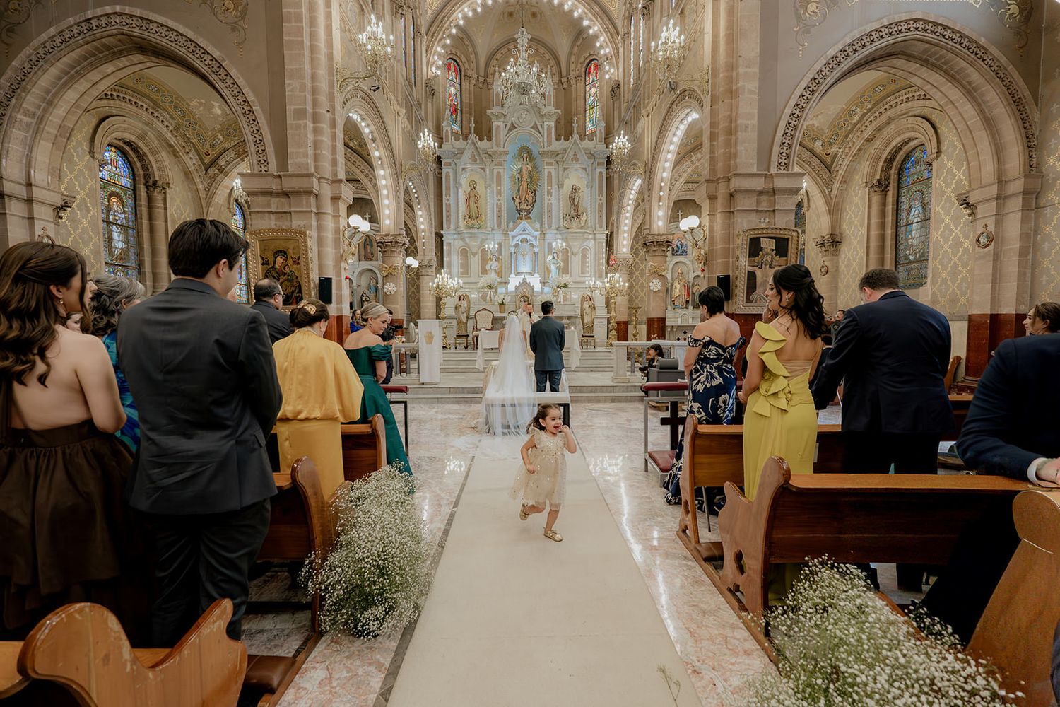 A flower girl walks down the aisle of an ornate church during a wedding ceremony with guests seated in wooden pews.