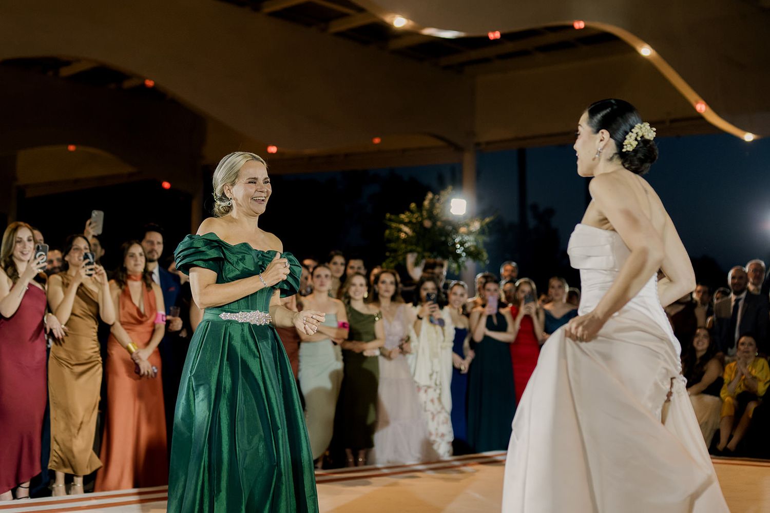 Bride in white gown shares a special dance moment with woman in emerald green dress at wedding.