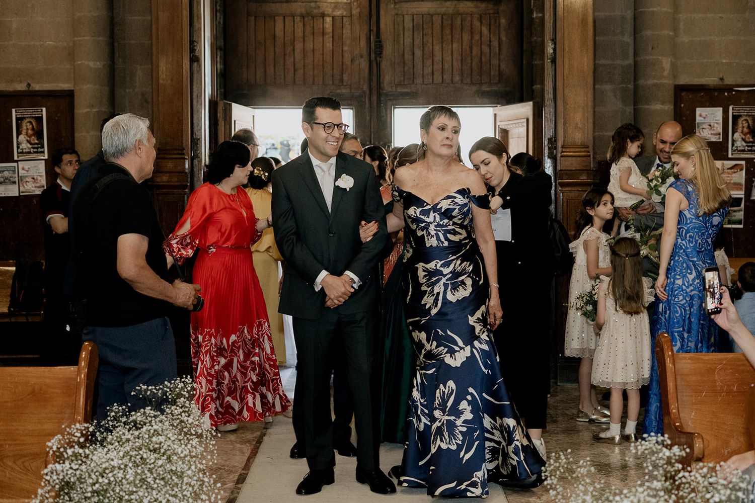 Groom in dark suit escorted down the church aisle by a woman in a floral gown during wedding ceremony.