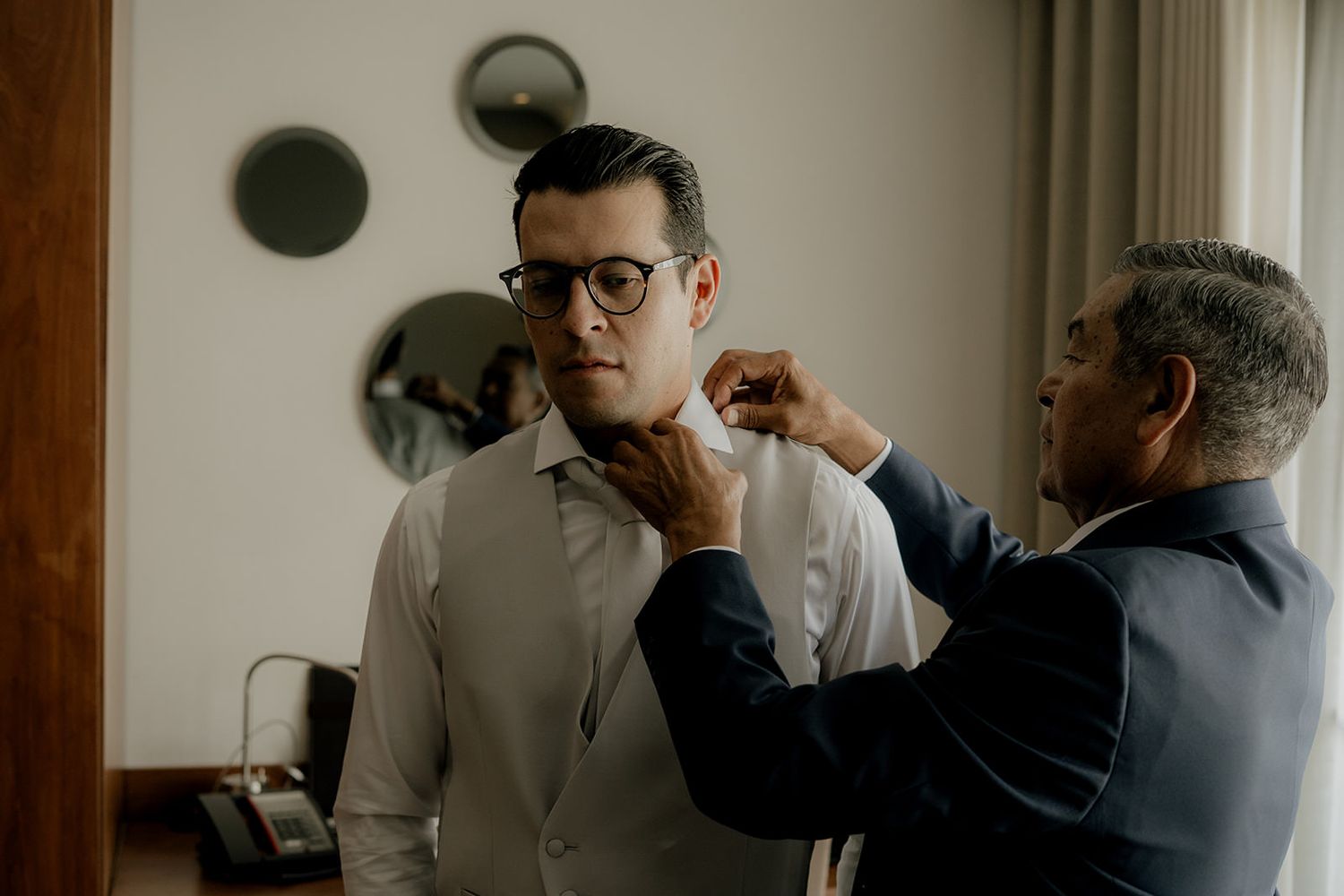 Groom in glasses having his bow tie adjusted by older man in hotel room before wedding.