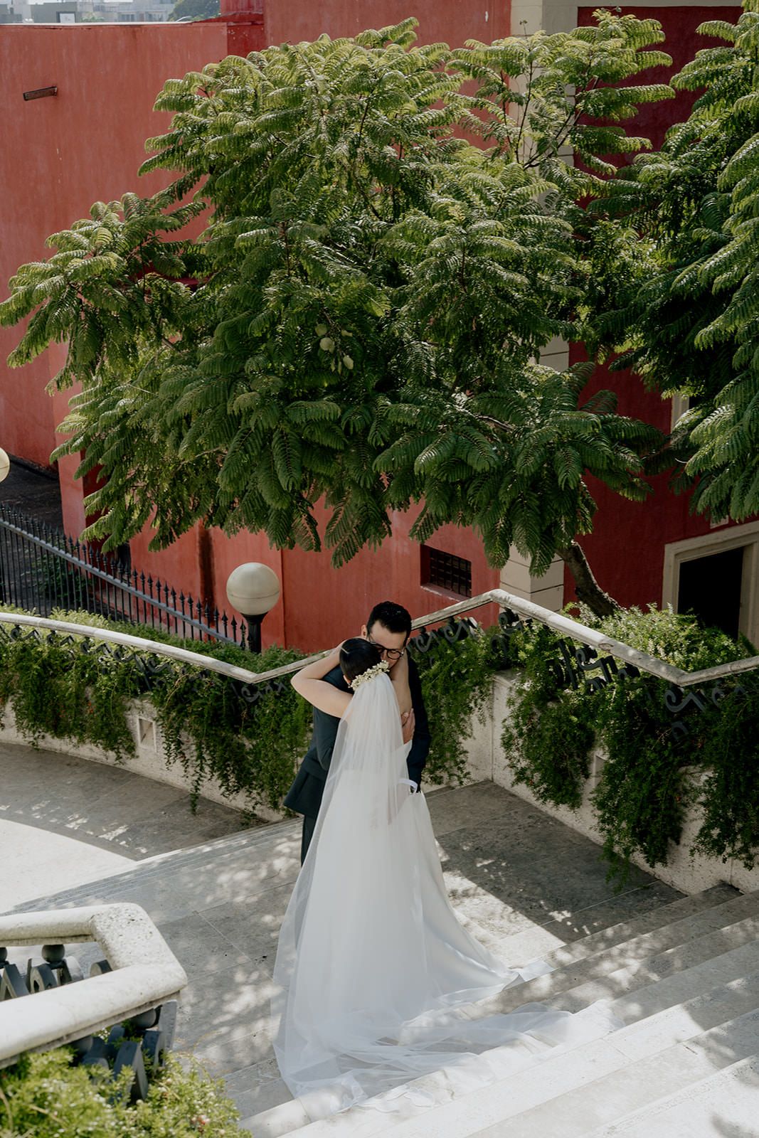 Bride and groom embracing on elegant staircase with lush green trees and red building backdrop.
