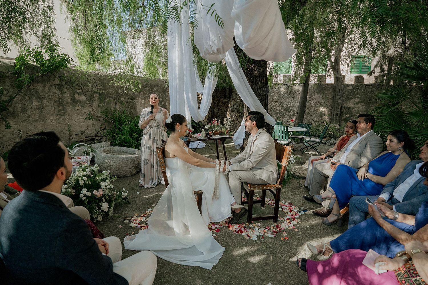 Intimate outdoor wedding ceremony under white drapes and willow trees with guests seated on both sides.