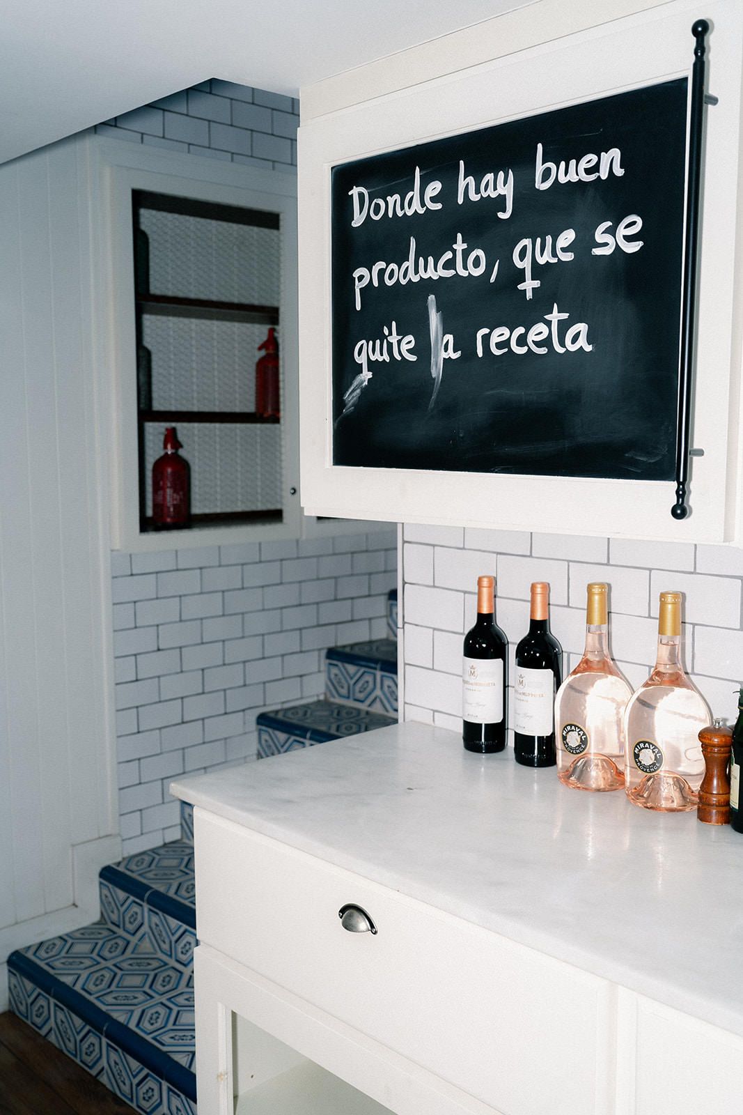 Modern white restaurant interior with Spanish text on black boards and wine bottles displayed on minimalist shelving.
