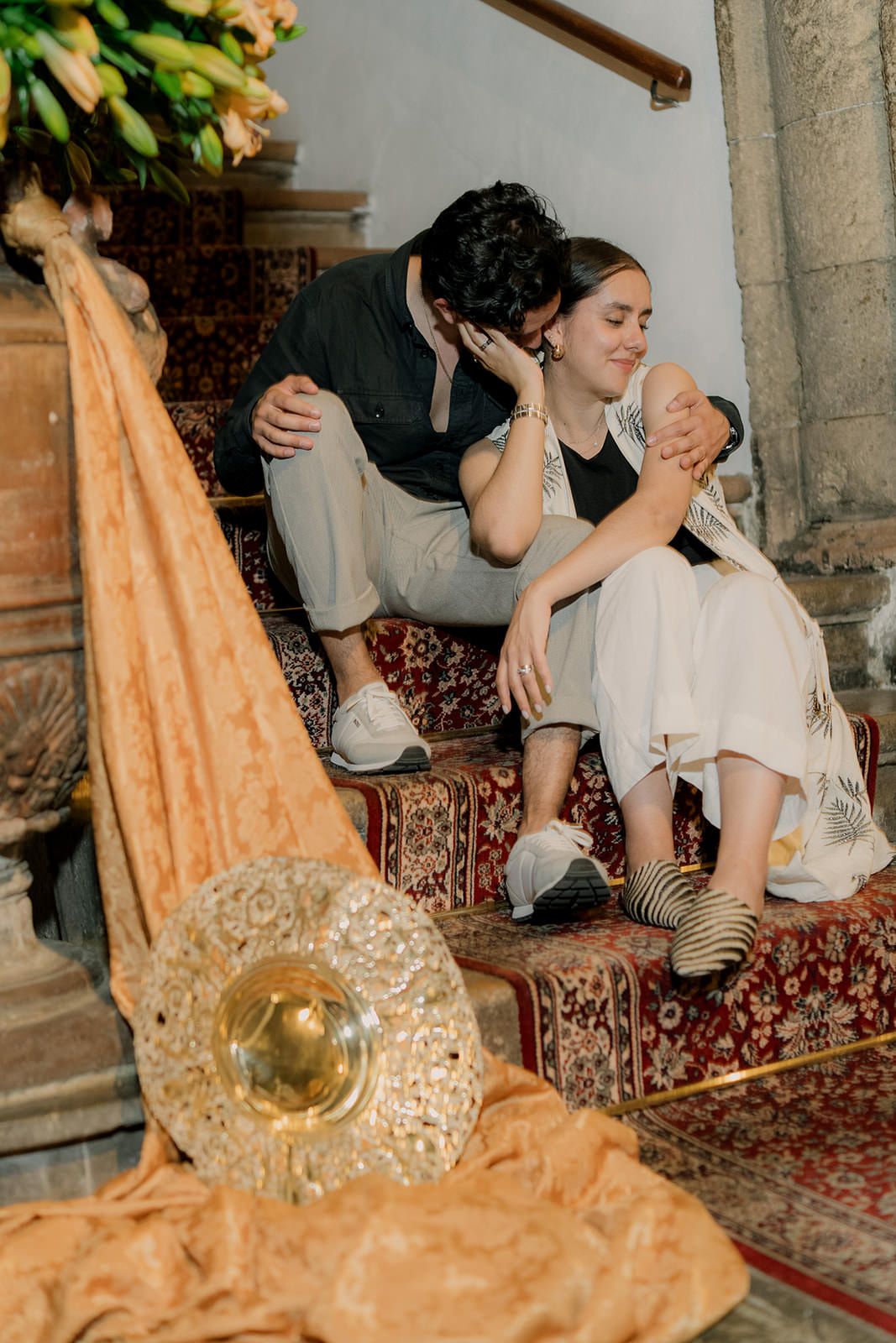 Intimate moment captured on ornate carpeted stairs with golden decor and peach-colored fabric.