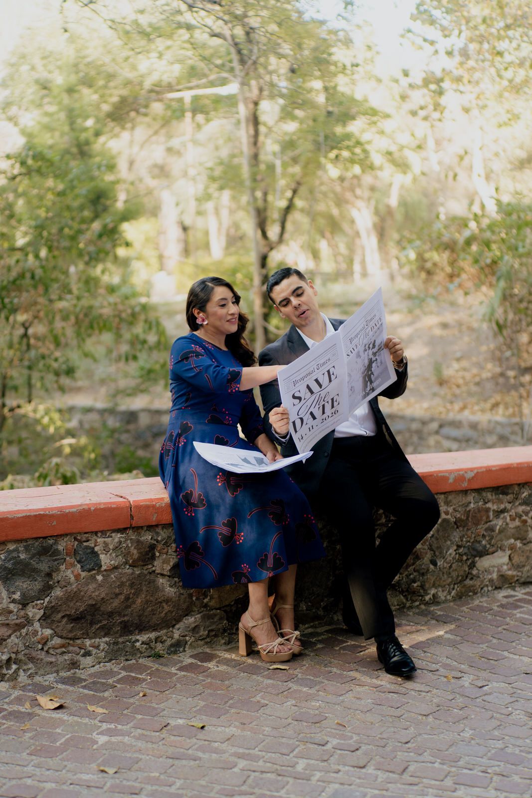 A couple sits on a stone wall sharing newspapers during their engagement session.