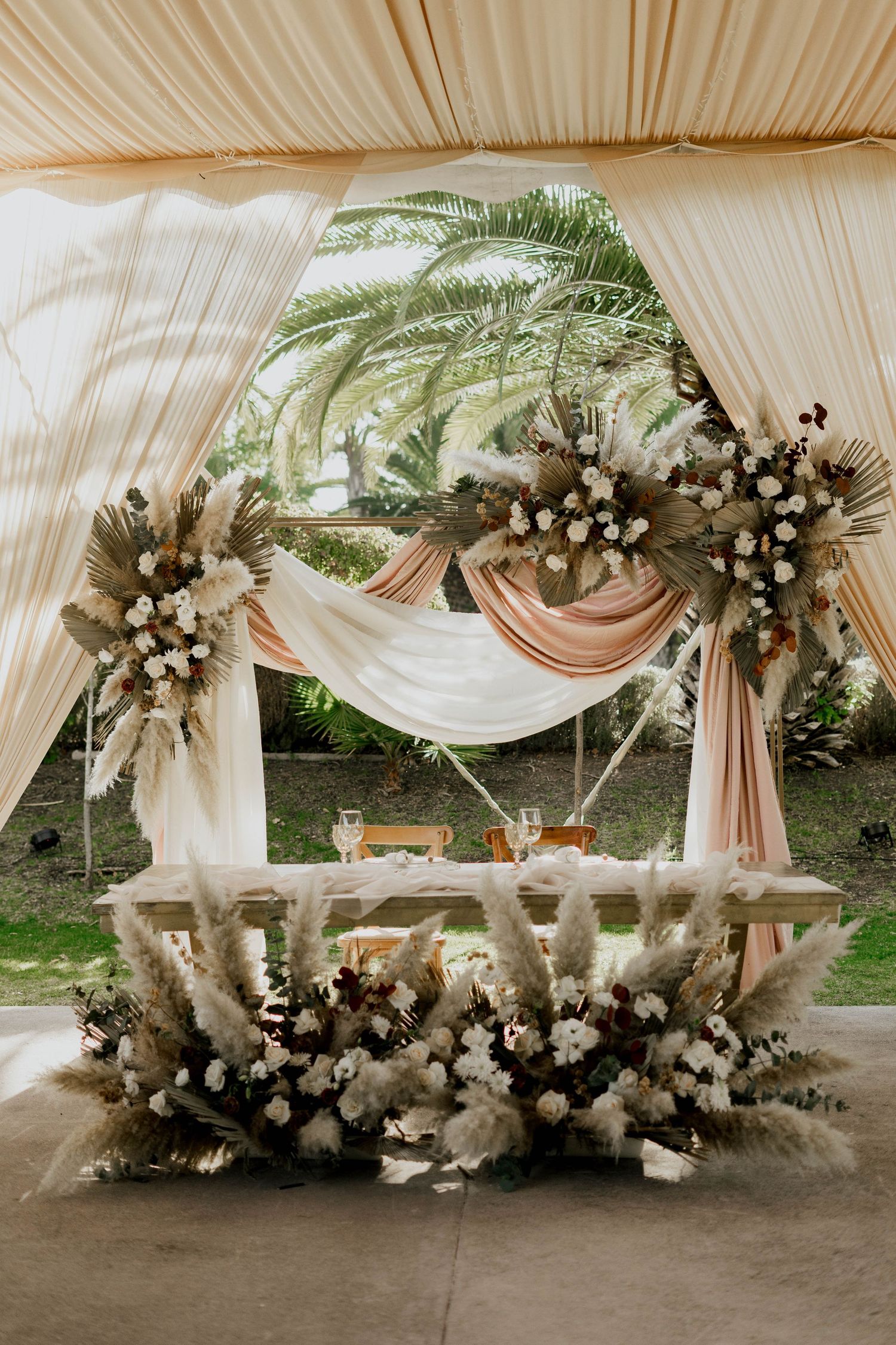 Romantic wedding arch decorated with white draping, pampas grass, and floral arrangements.