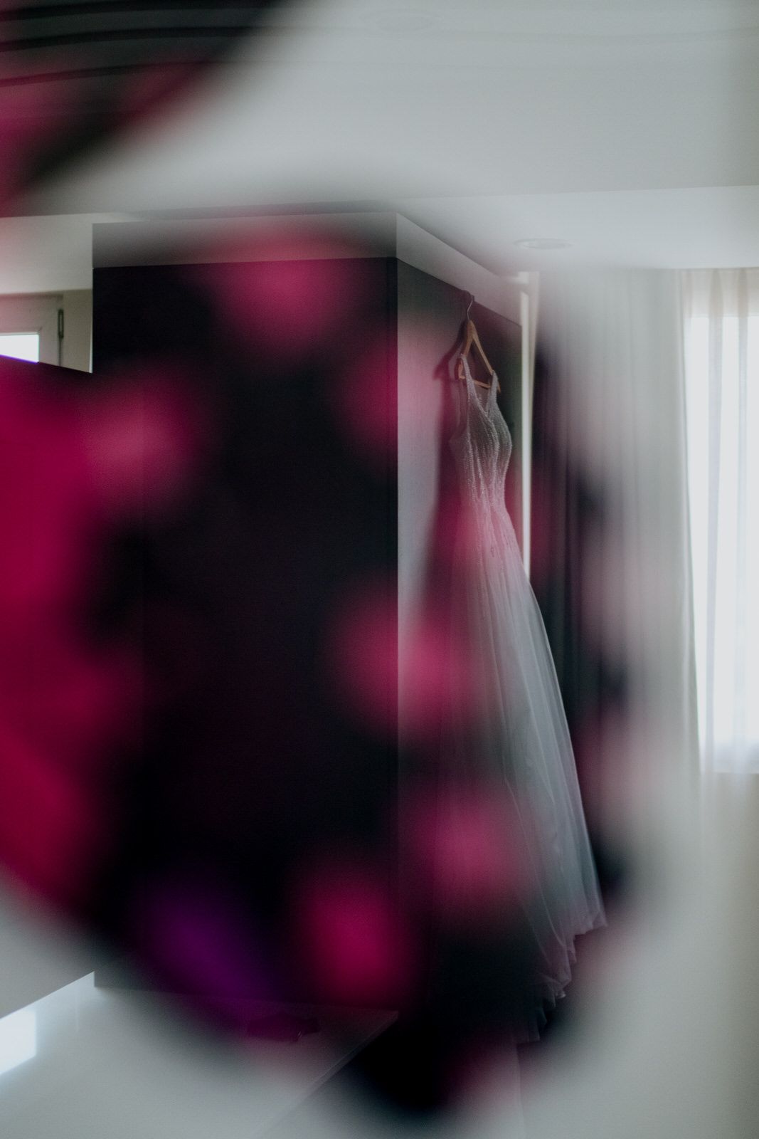 A white wedding dress hangs elegantly in a softly lit room, framed by blurred pink floral bokeh in the foreground.