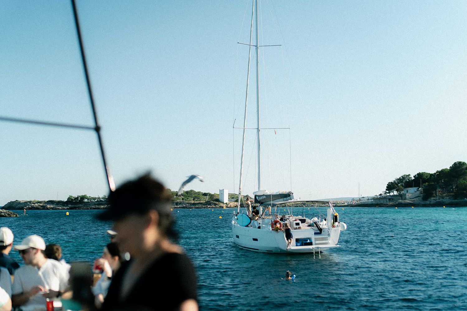 Sailboats anchored in a tranquil blue bay with scattered boats and people enjoying the water on a sunny day.