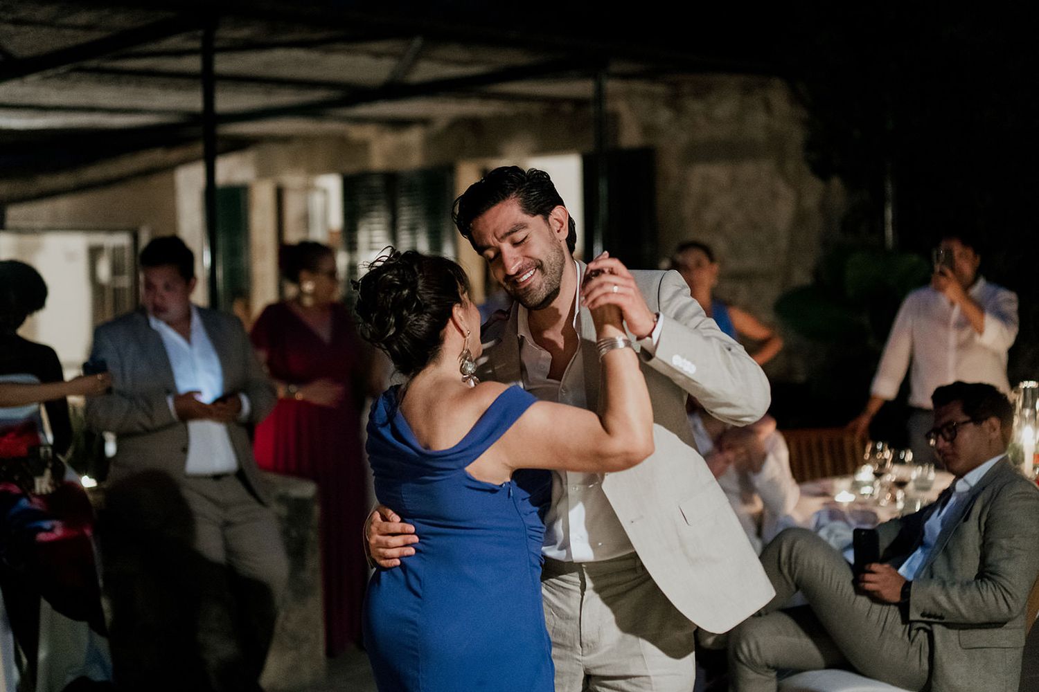 A couple shares an intimate dance at an indoor reception venue with guests looking on in the background.