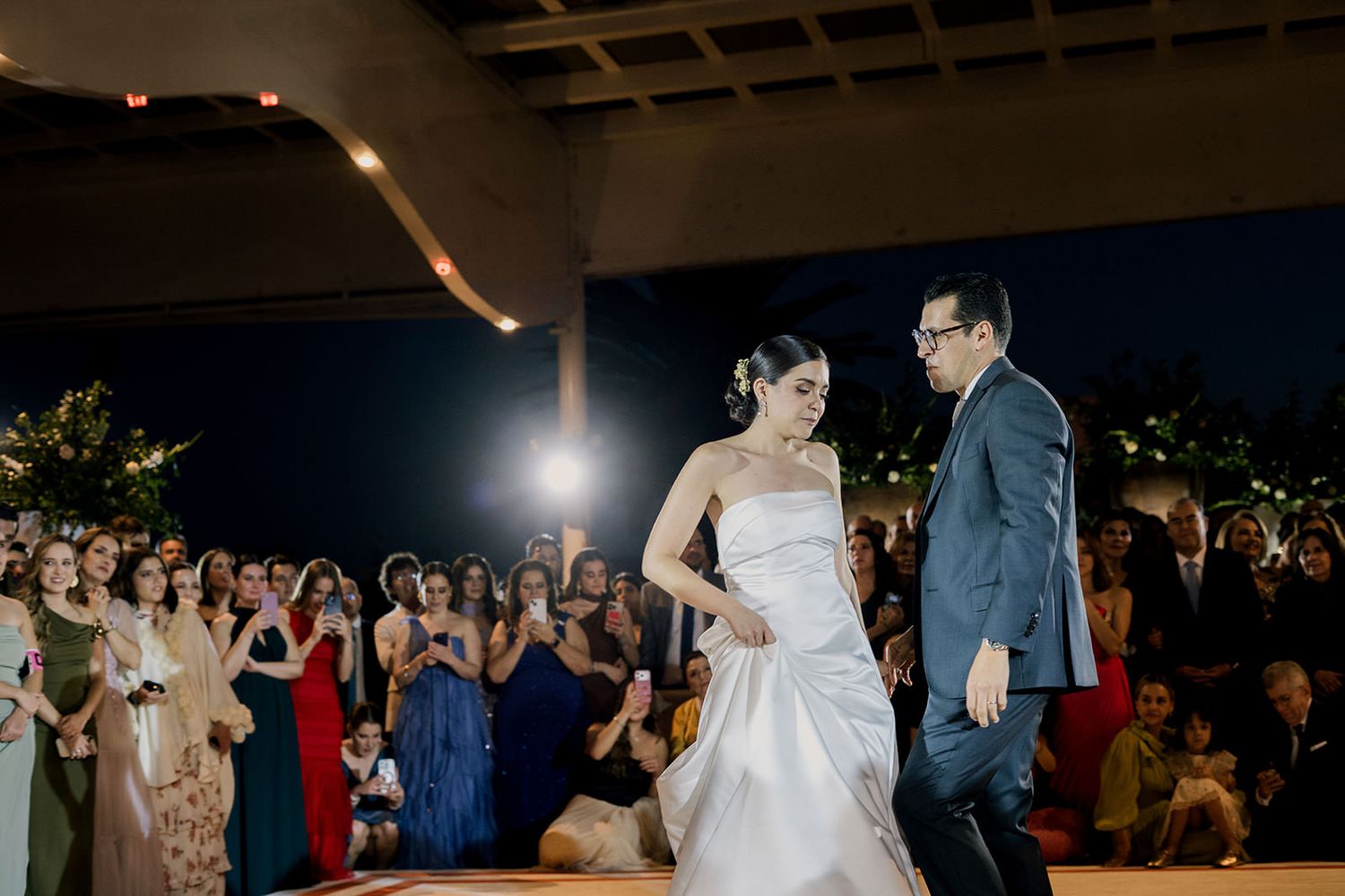 Bride in white gown and groom in navy suit enter wedding reception to applause from elegantly dressed guests.