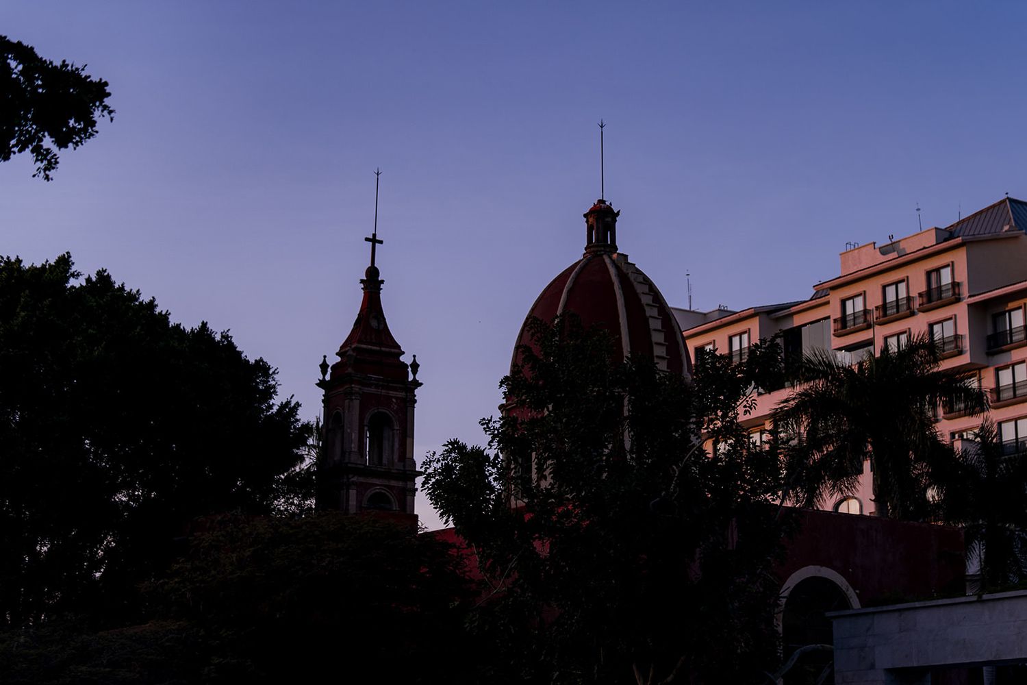 Silhouette of historic church dome and bell tower against purple dusk sky with trees.