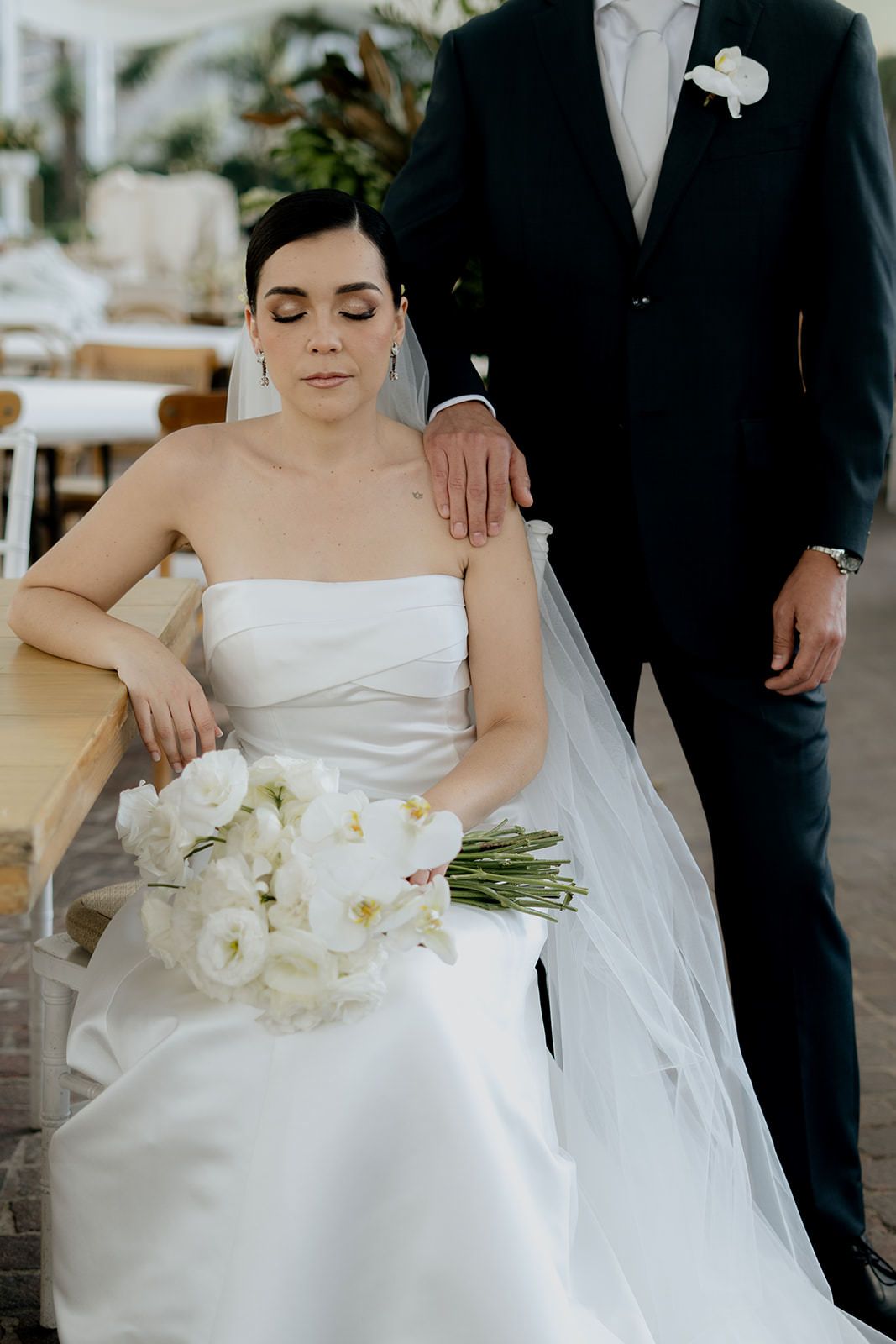 Elegant bride in strapless white gown holds bouquet of white flowers while groom in dark suit stands beside her.