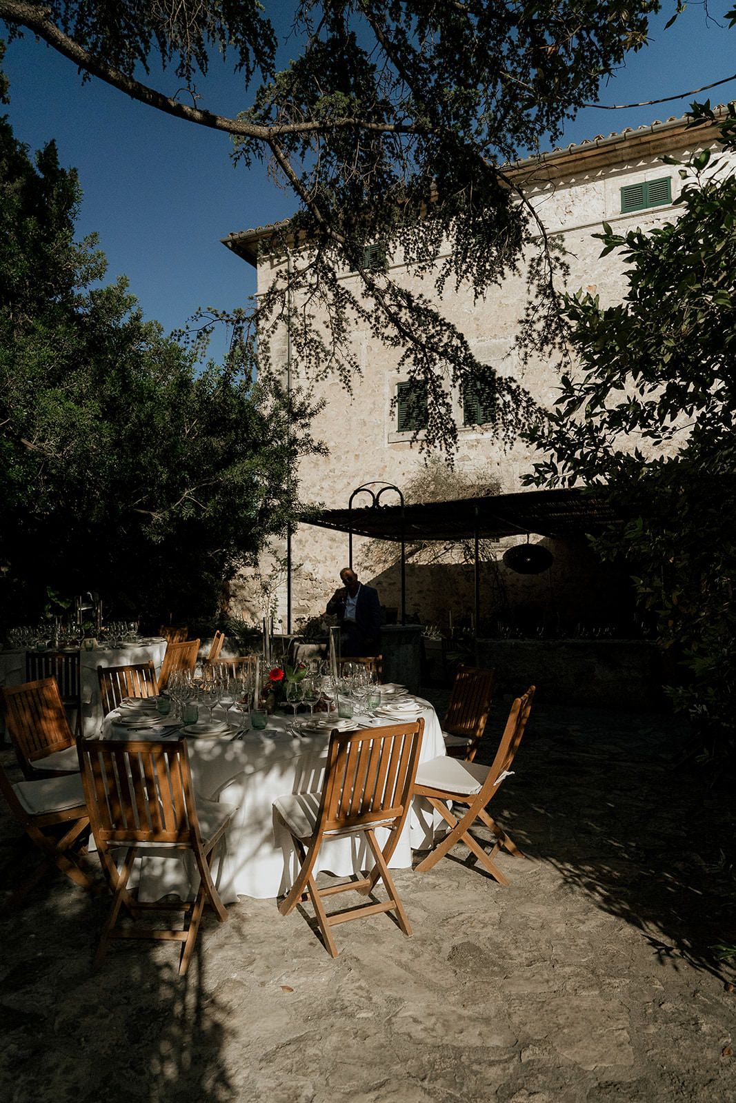 Wooden folding chairs arranged on a stone patio with white linens under shady trees.