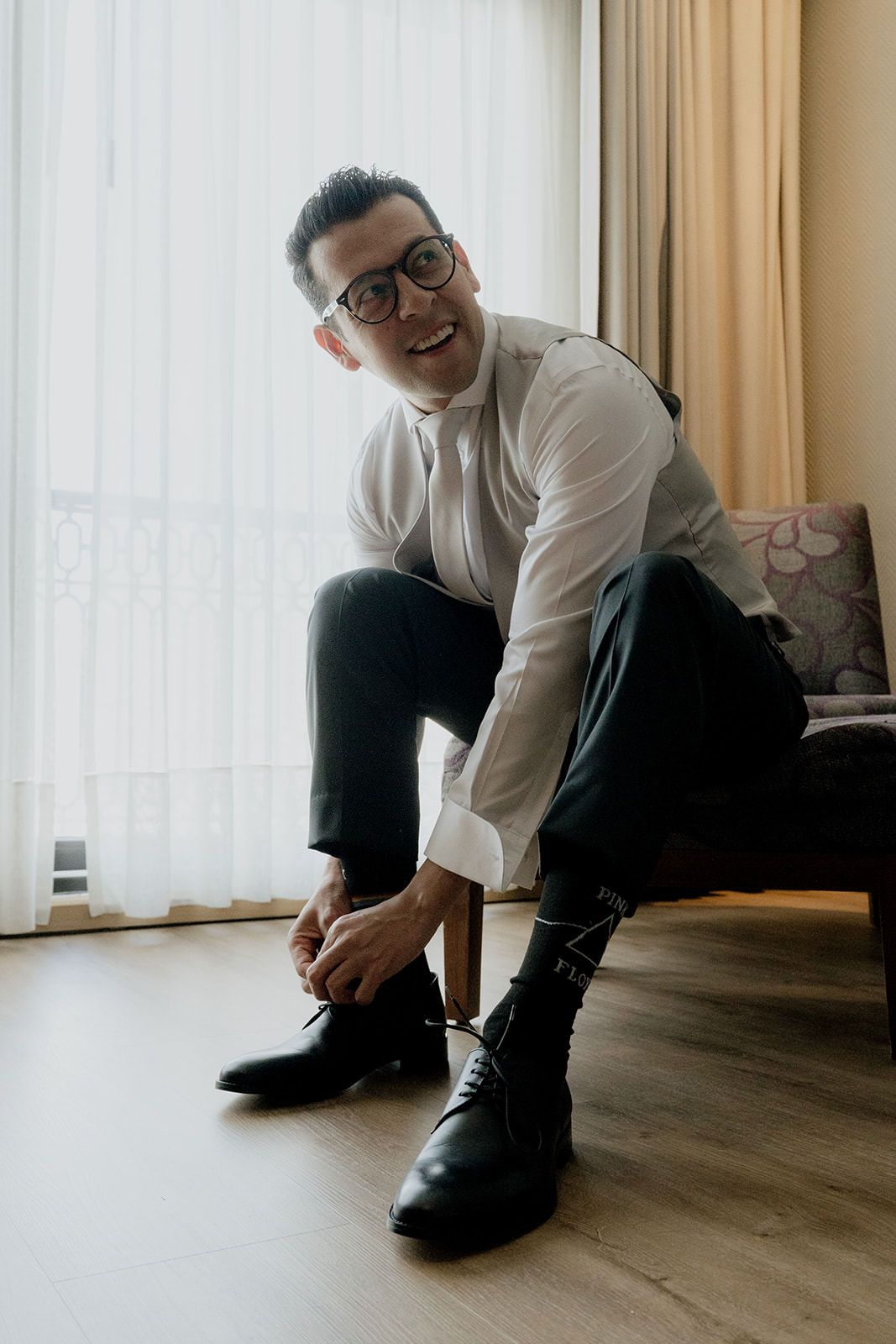 Smiling groom in white dress shirt and glasses tying black dress shoes before wedding ceremony.