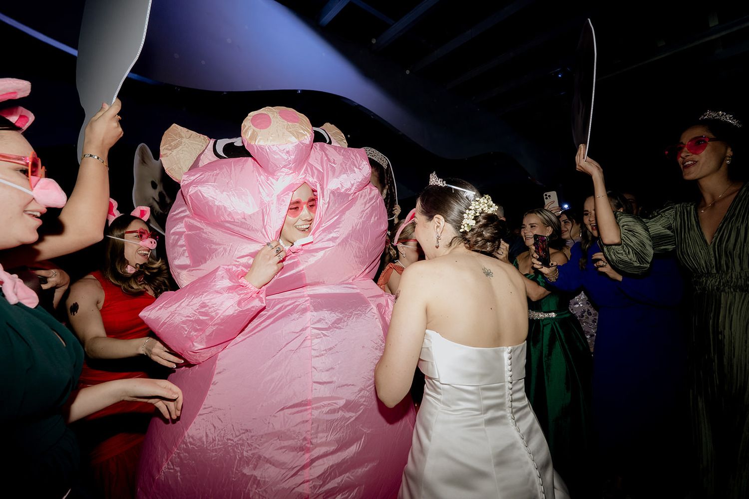 Bride in white dress dances with guest in pink inflatable pig costume at lively wedding reception party.