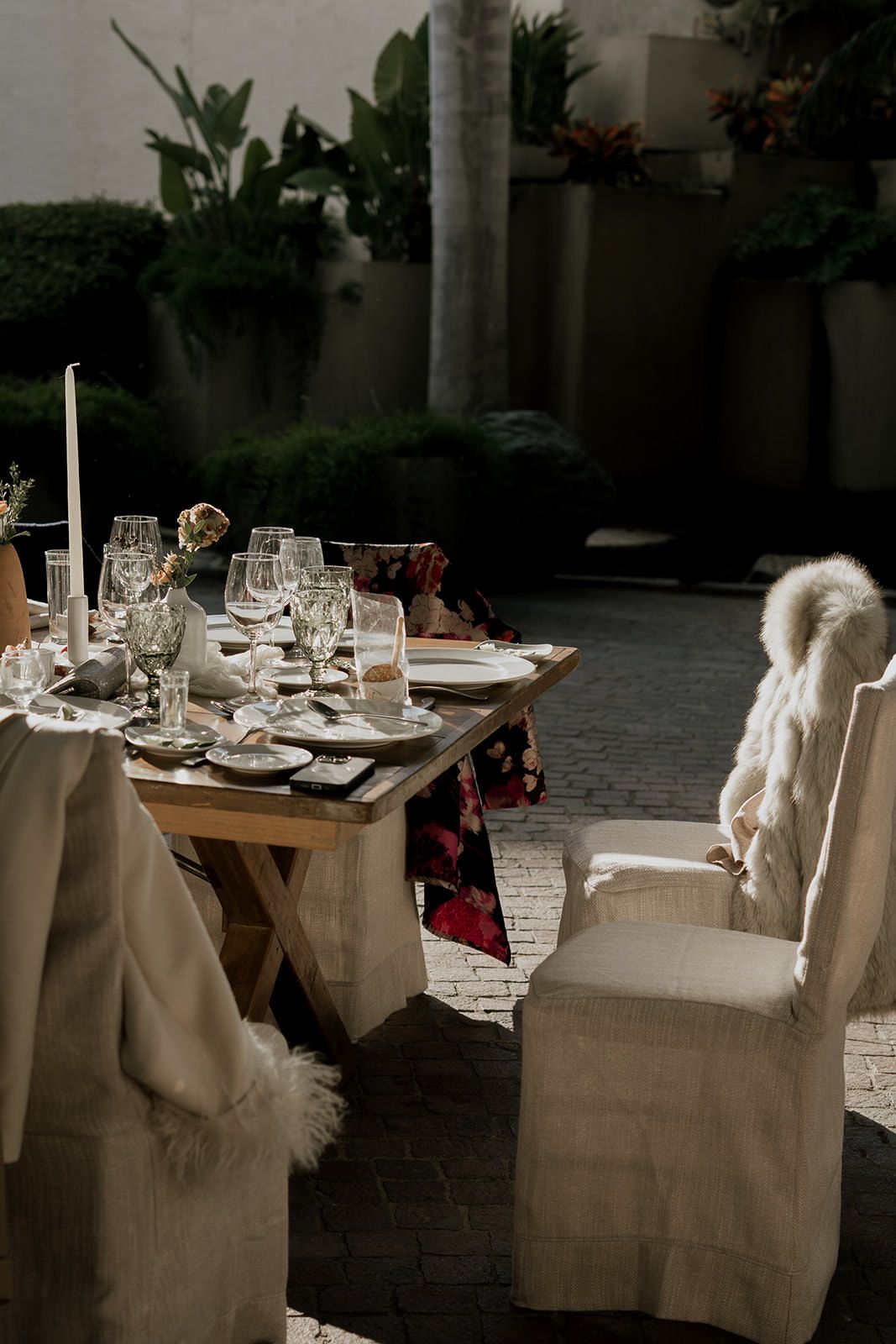 Elegant outdoor wedding reception table set with glassware, candles, and floral decor, surrounded by white linen chairs with fur throws.