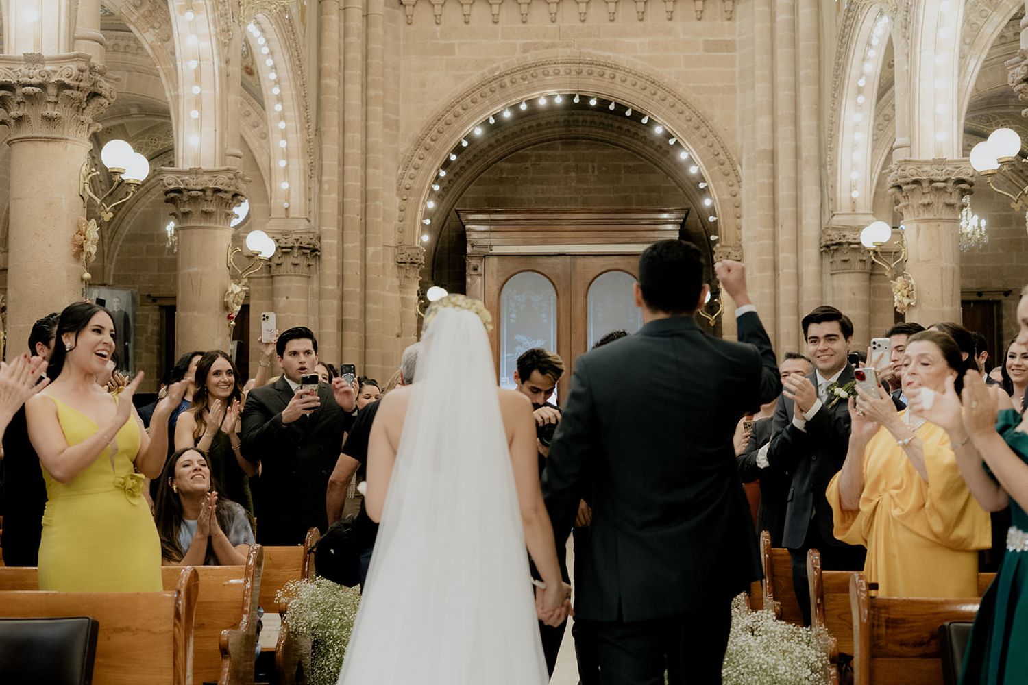 Bride and groom walk down the aisle of an ornate church as guests applaud during a wedding ceremony.