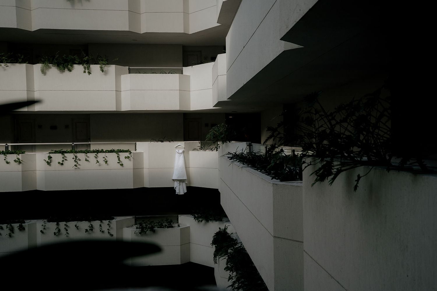 White wedding dress hanging on a modern multi-story atrium balcony surrounded by lush green plants.