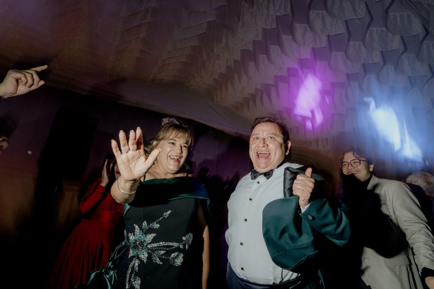 Joyful wedding guests dancing and celebrating under decorative ceiling with colorful purple disco lights.