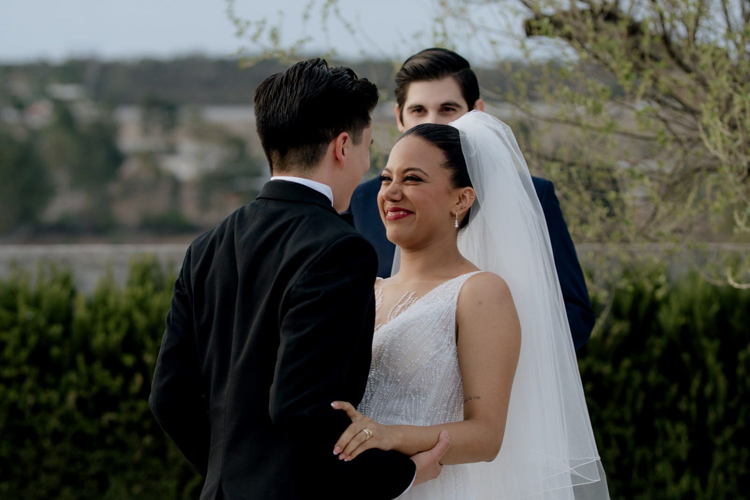 Smiling bride in white veil holds grooms hands during outdoor wedding ceremony.