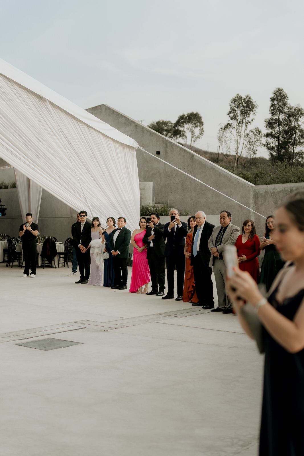 Wedding guests in formal attire line up outdoors beside white draped tent at elegant venue.