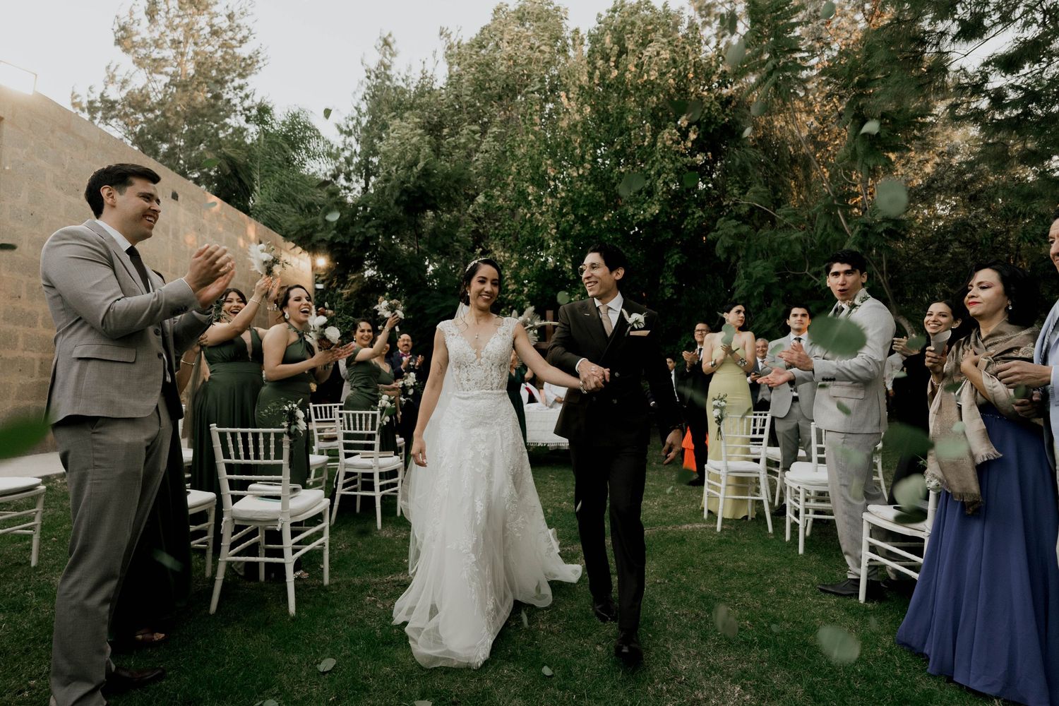 Garden wedding reception with guests gathered among white chairs and natural greenery.