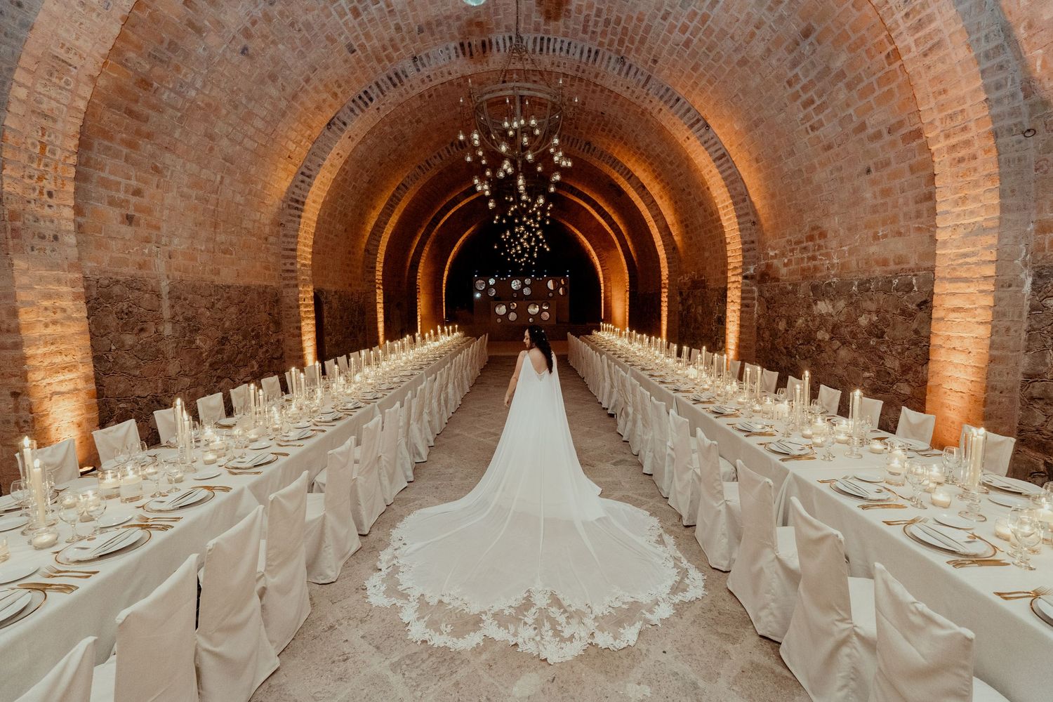 Long banquet tables arranged in an arched brick cellar with white linens and candlelight.