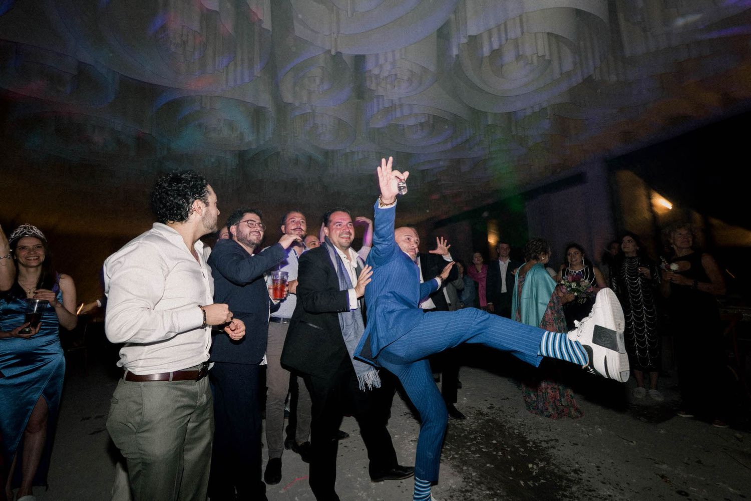 Wedding guests dance energetically on the reception dance floor under dramatic lighting.