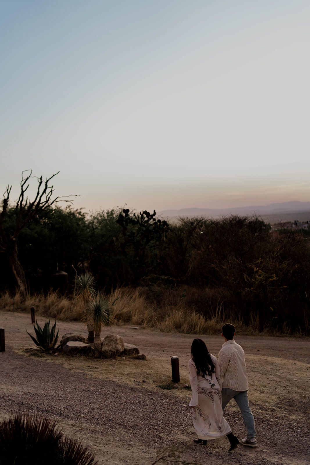 A couple walks together on a desert path during sunset with mountains in the background.