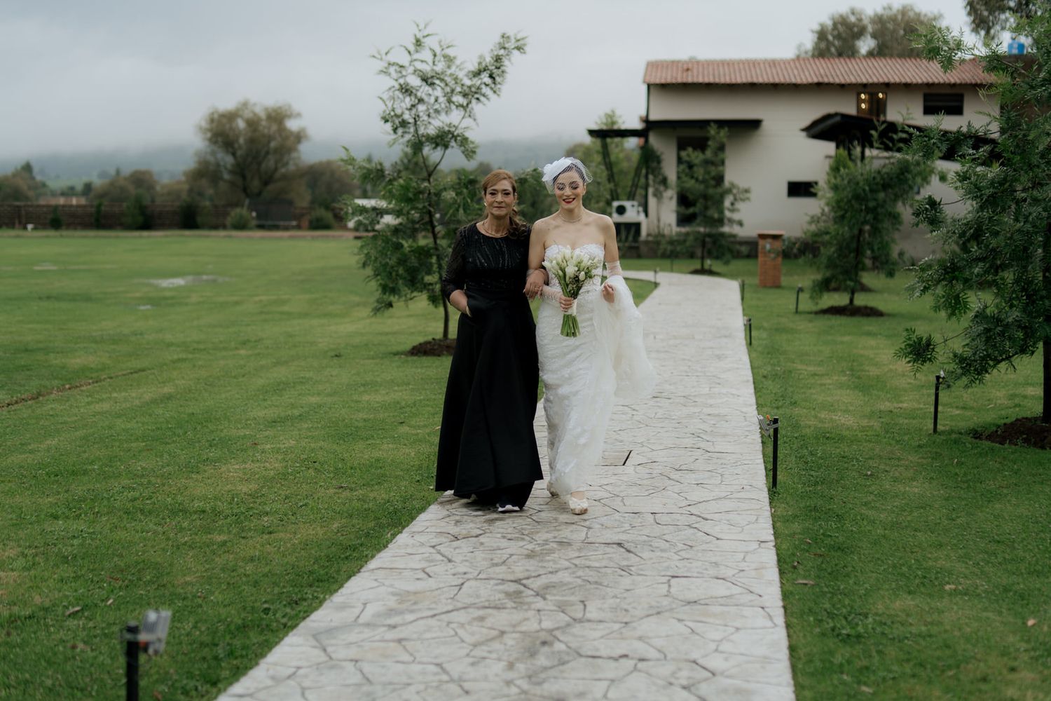 Wedding couple walks along a scenic outdoor path surrounded by green lawns and trees.