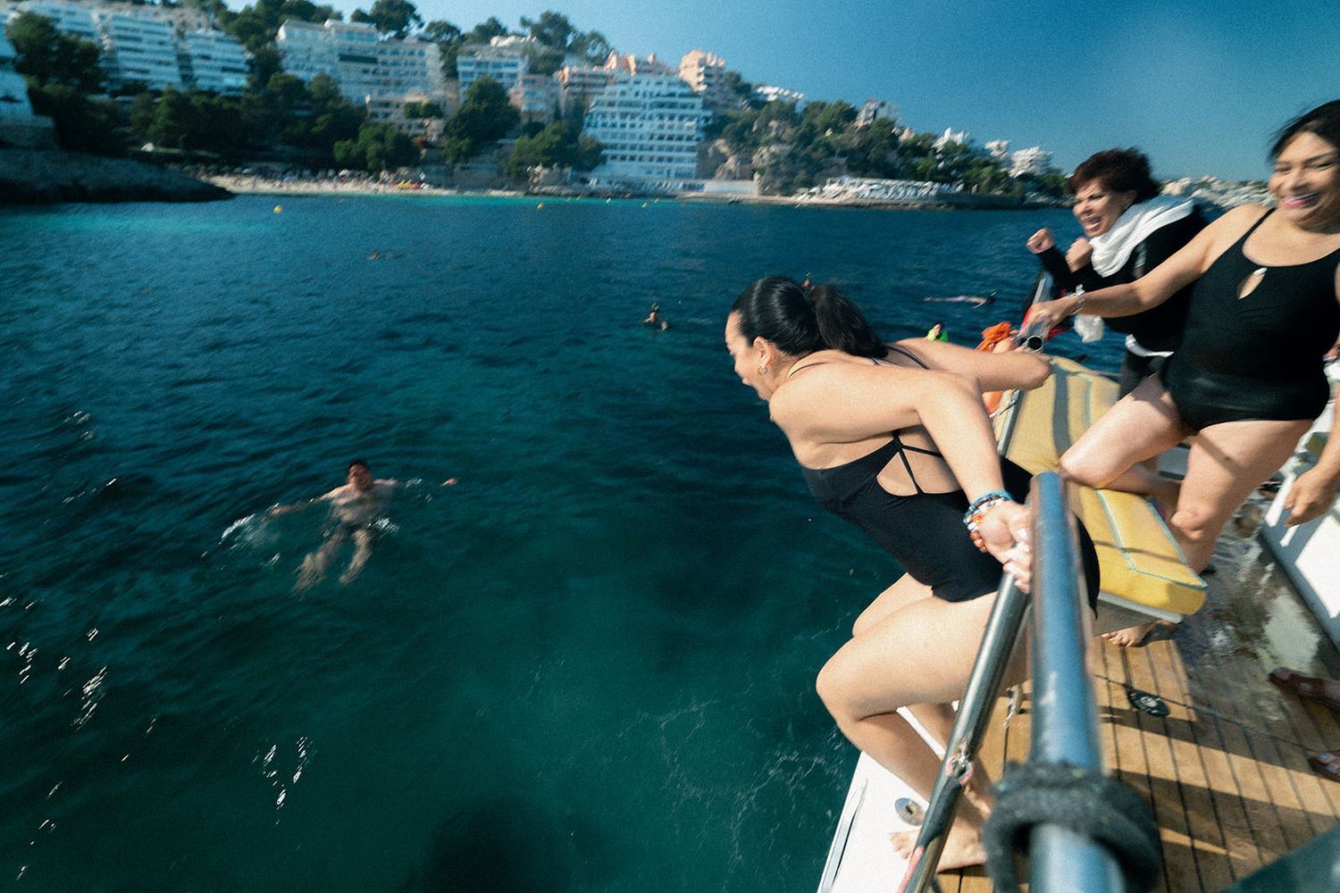 People jumping into clear turquoise Mediterranean waters from a wooden deck platform near coastal buildings.