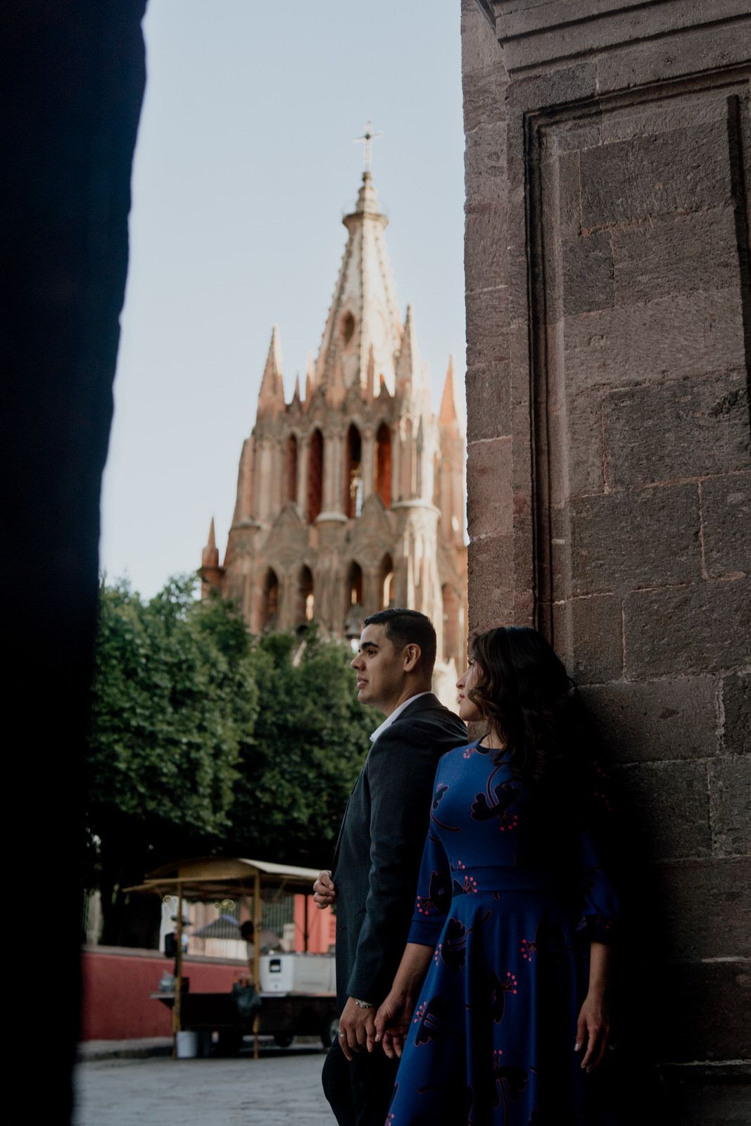 A couple poses against a stone wall with gothic church spires visible in the background.