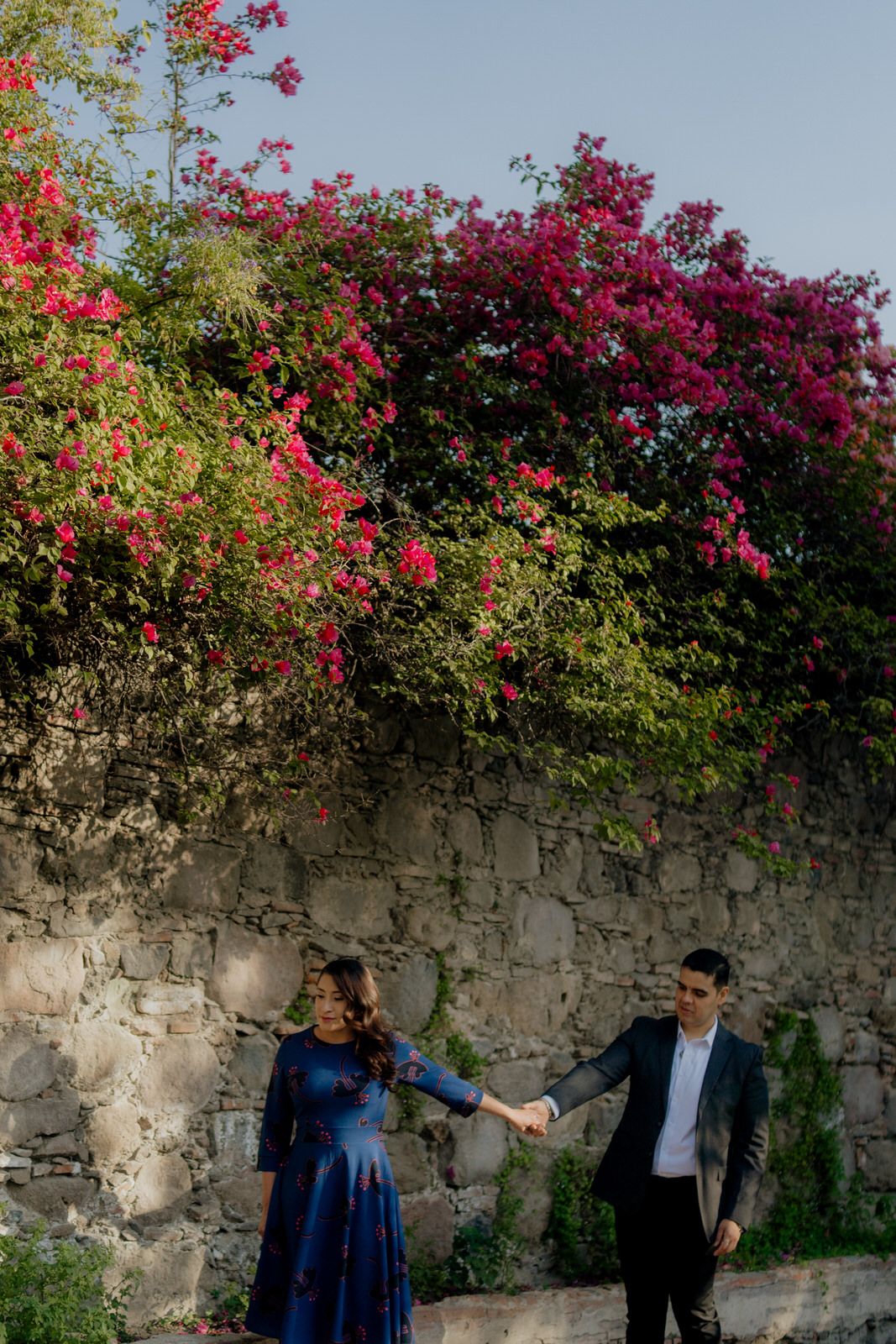 Stone wall adorned with vibrant pink bougainvillea flowers against blue sky.