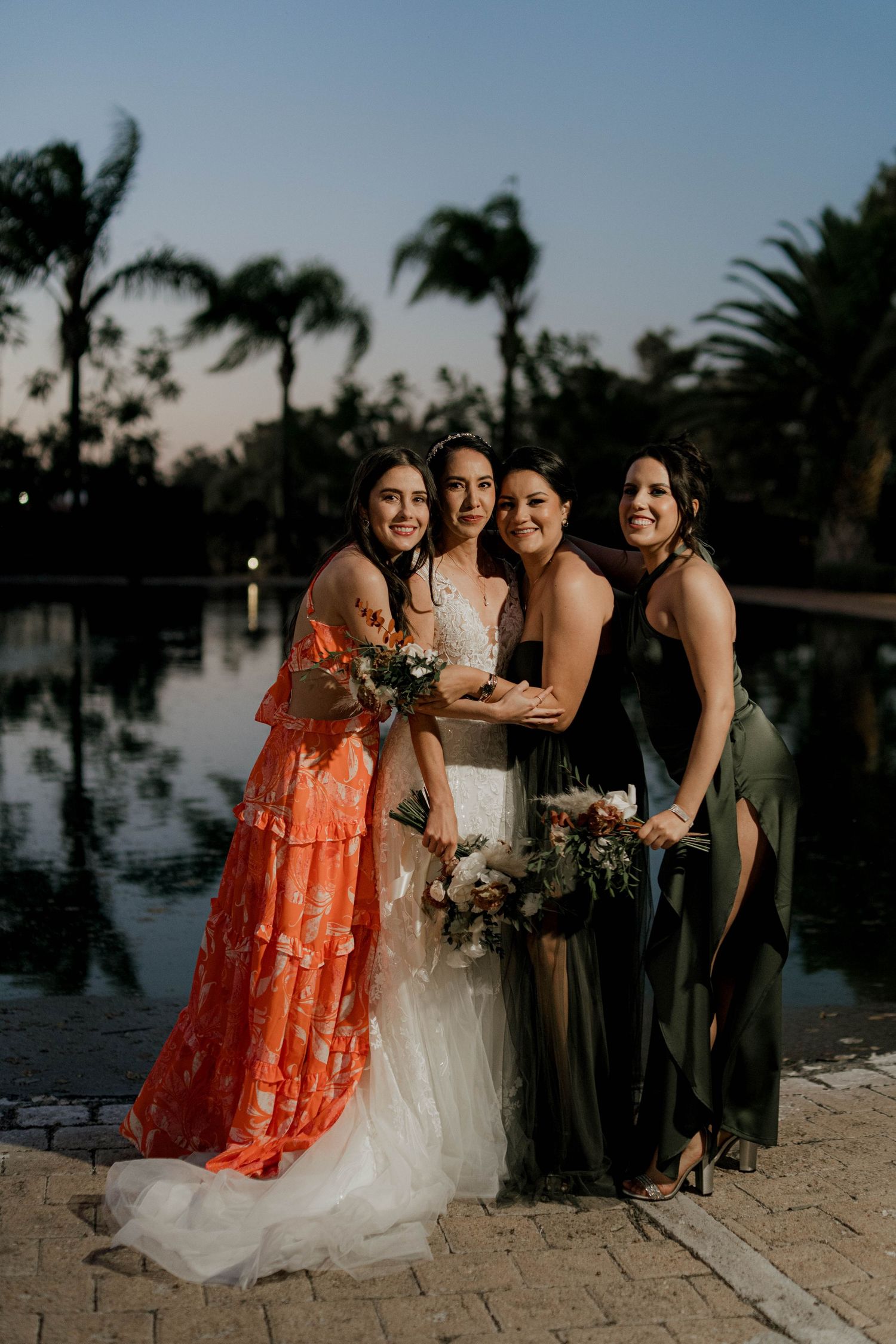 Bridal party in coral and olive dresses pose together at sunset by the water.