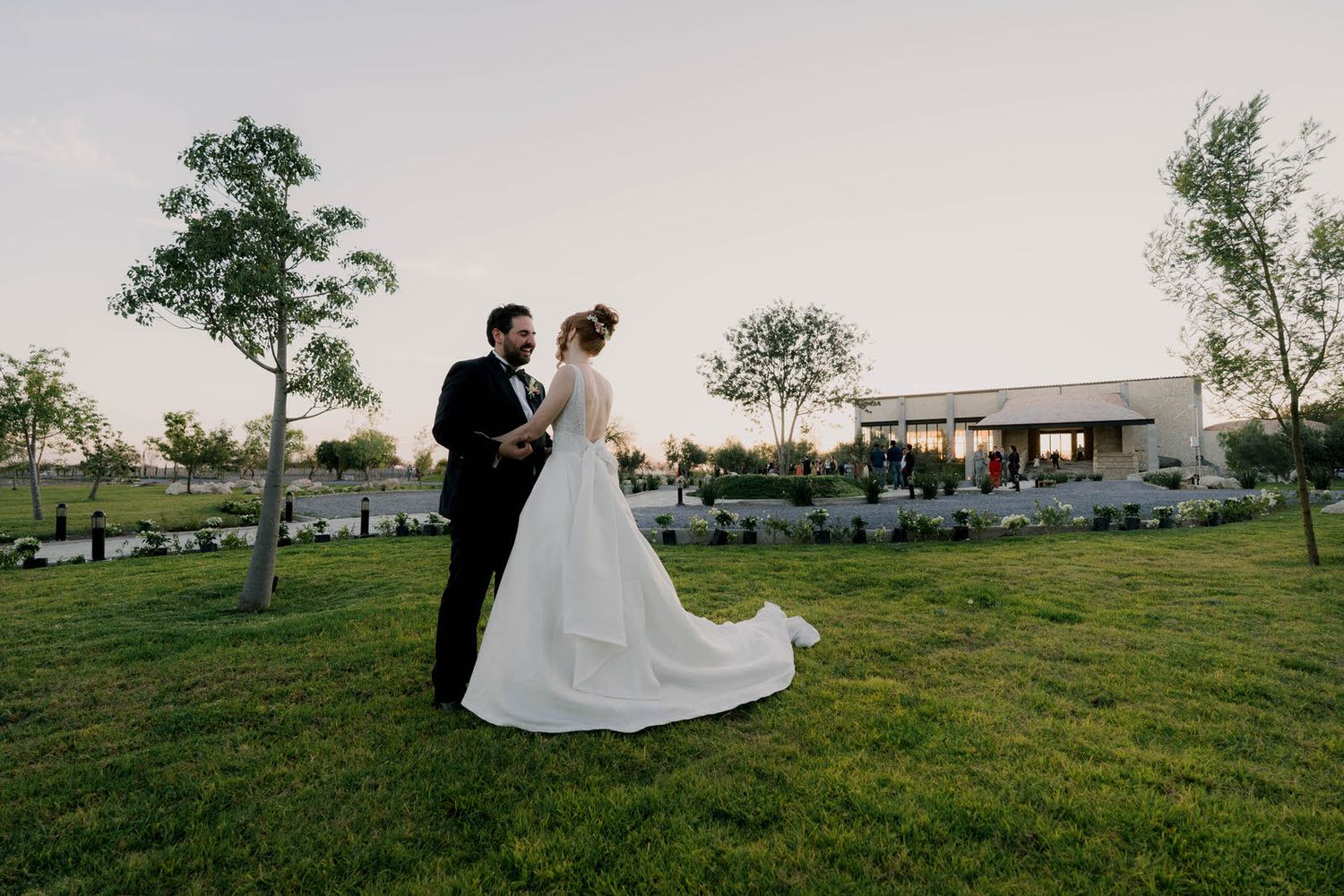 A wedding couple stands together on a grassy hill overlooking a scenic venue at dusk.