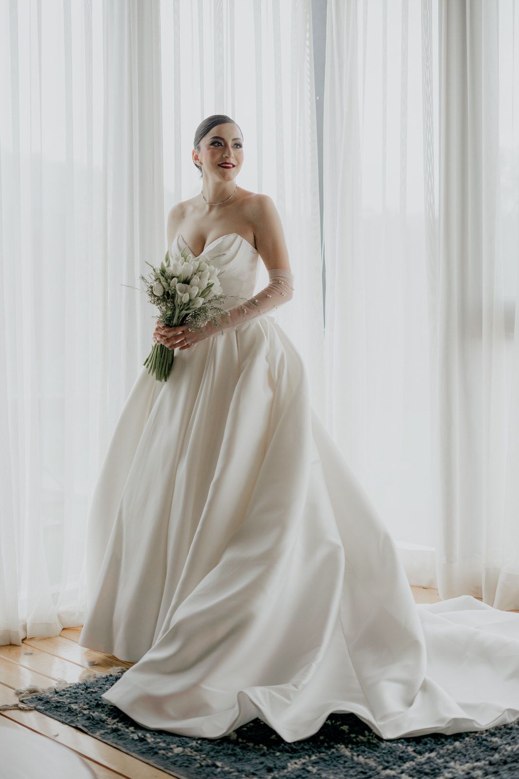 Elegant wedding portrait featuring a strapless ballgown and simple bouquet by sheer curtains.